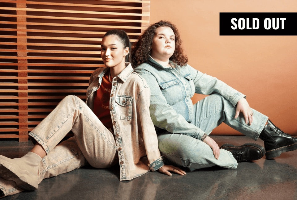 Two models sit back to back on floor in front of wood paneled backdrop. They wear denim pieces from the Nordstrom BP x Wildfang collection. A banner in the top right corner reads SOLD OUT.