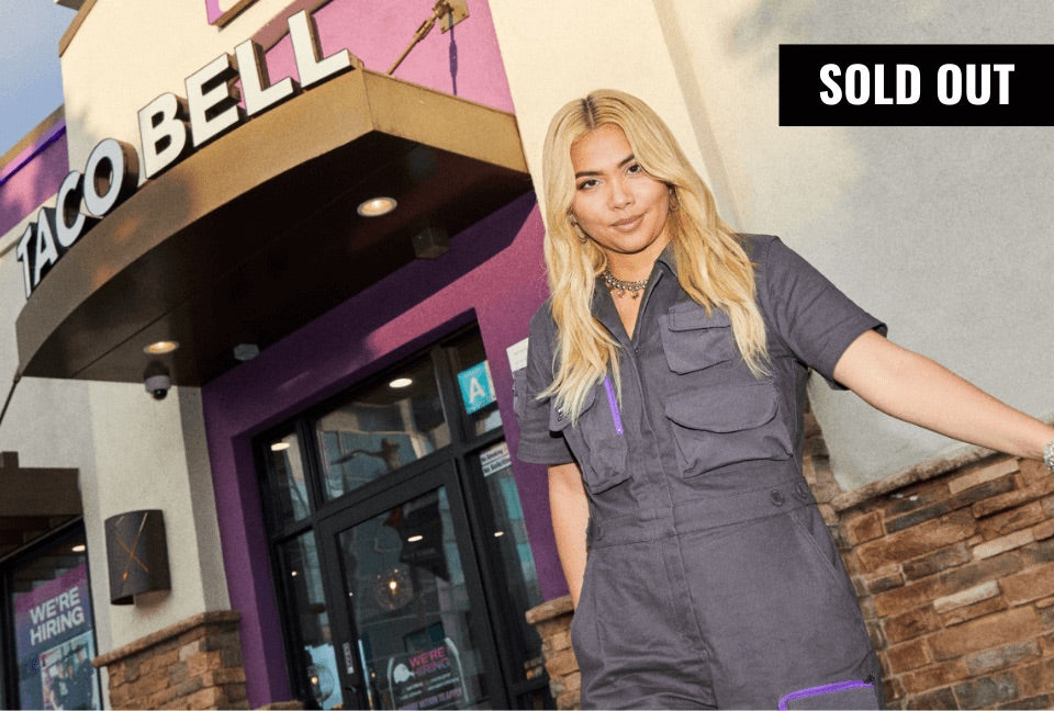 Hayley Kiyoko stands in front of entrance to Taco Bell looking at camera wearing The Taco Bell x Wildfang Coverall. A banner in the top right corner says SOLD OUT.