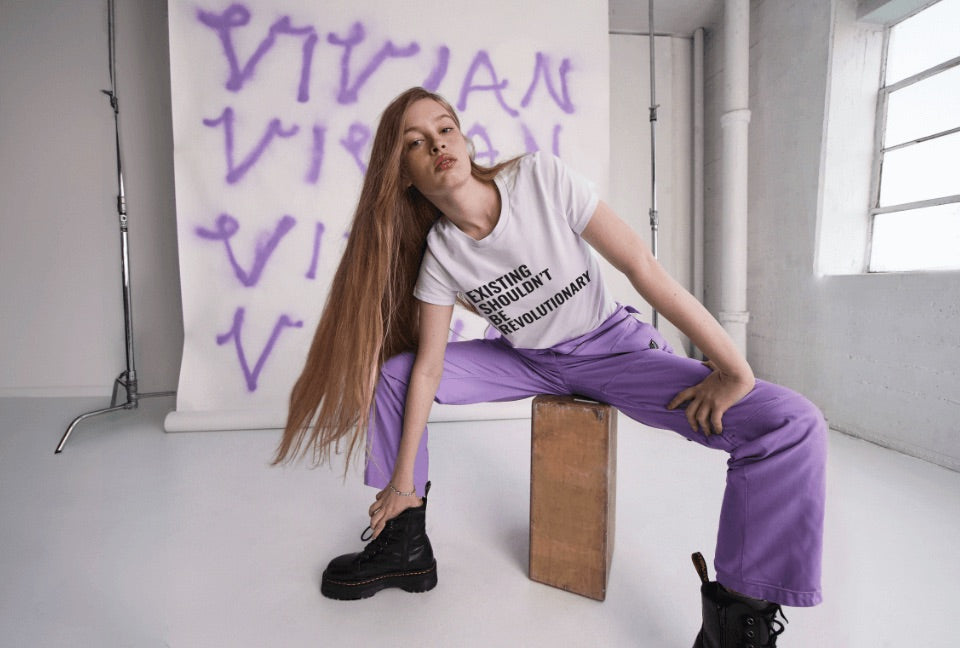 Vivian Wilson sits on wood block leaning over with hand on ankle, her blonde hair draped over her shoulder, wearing The Essential High Waisted Coverall in Lilac, the Vivian Wilson for Wildfang Tee, and Doc Marten black boots. Behind her, a white backdrop features her name spray painted in purple multiple times. 