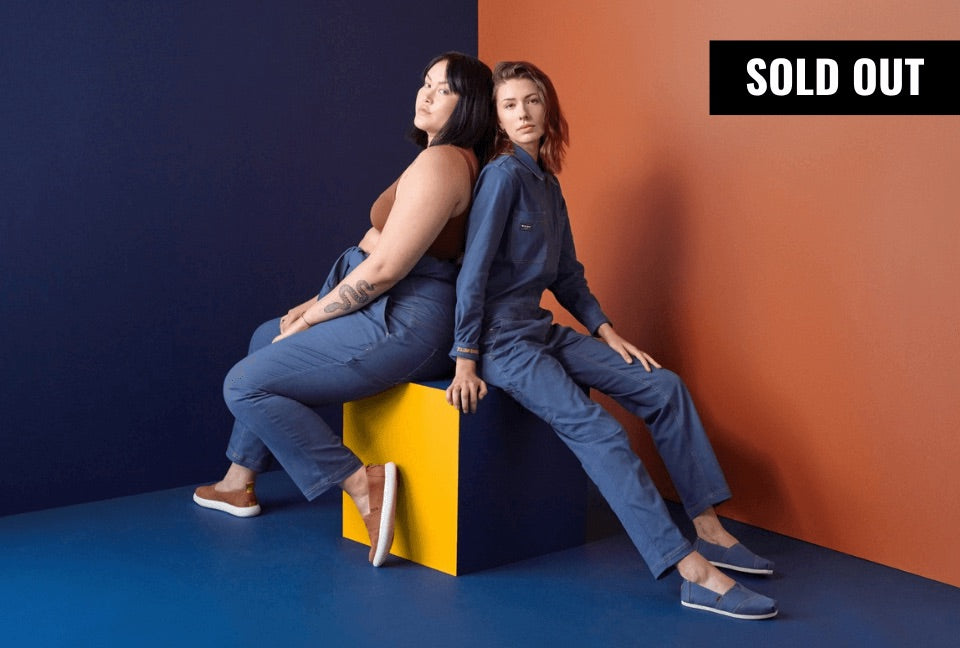 Two people sit back to back on top of blue and yellow cube in front of blue and orange background. They wear blue coveralls and Toms shoes. A banner in the top right corner reads SOLD OUT.