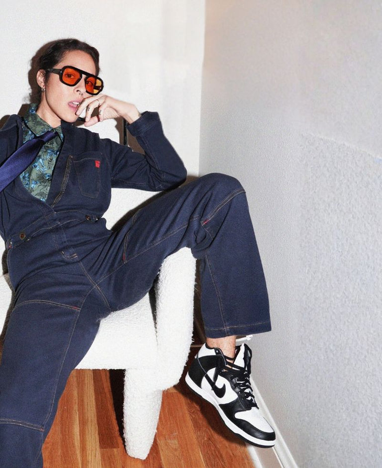 Person sits in white chair wearing blue denim coverall and sneakers.