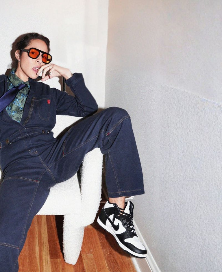 Person wearing denim coverall sits in white chair with one leg over chair arm in room with white walls