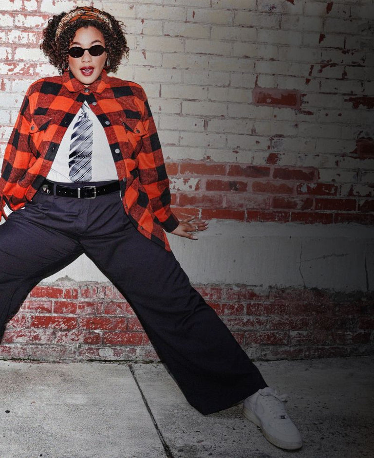 Person leans against brick wall wearing plaid jacket, black pants, and white sneakers.