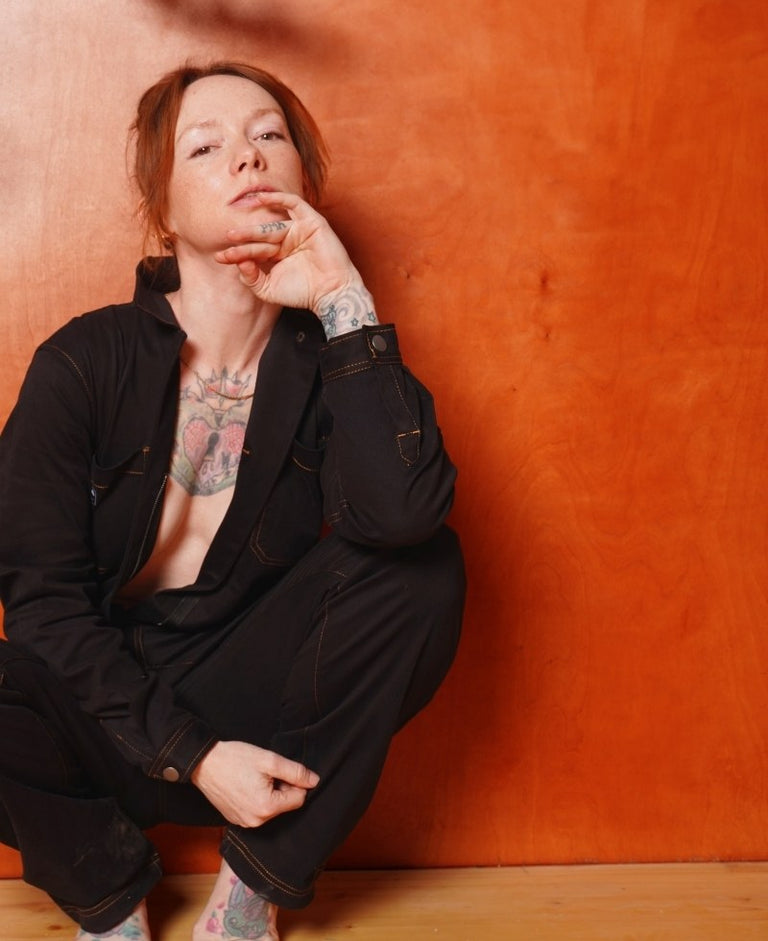 Against orange toned wood background, model wearing black long sleeve coverall squats down while looking at camera.