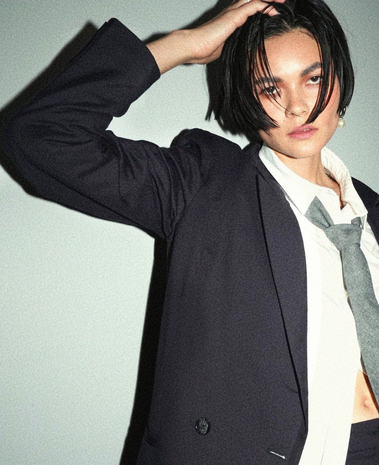 Close up image of person with short black hair wearing black suit, white button up, and grey tie, standing against grey wall.
