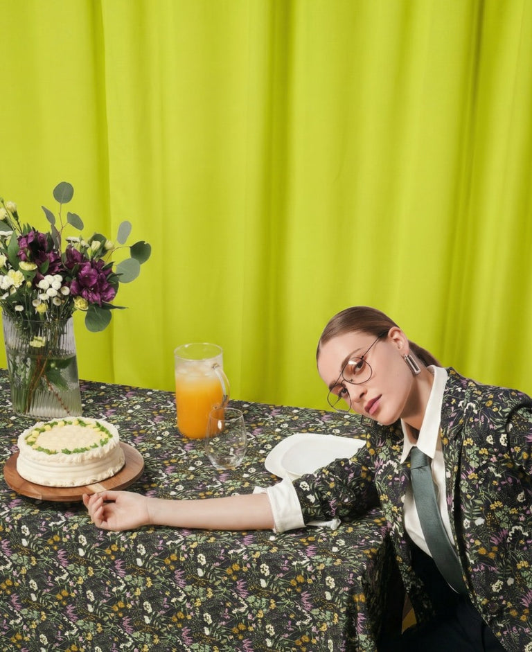 Person wearing floral suit sits by table with matching tablecloth against yellow curtain background.