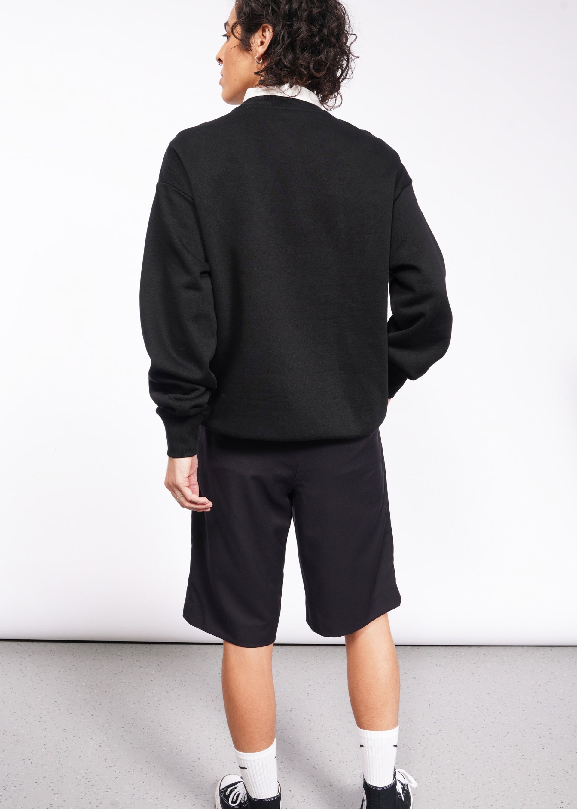 A person with curly hair stands against a plain background, dressed in a black Wild Feminist Fleece Crew, black shorts, white socks, and black sneakers. The brushed fleece adds comfort while subtly capturing the spirit of a WILD FEMINIST.