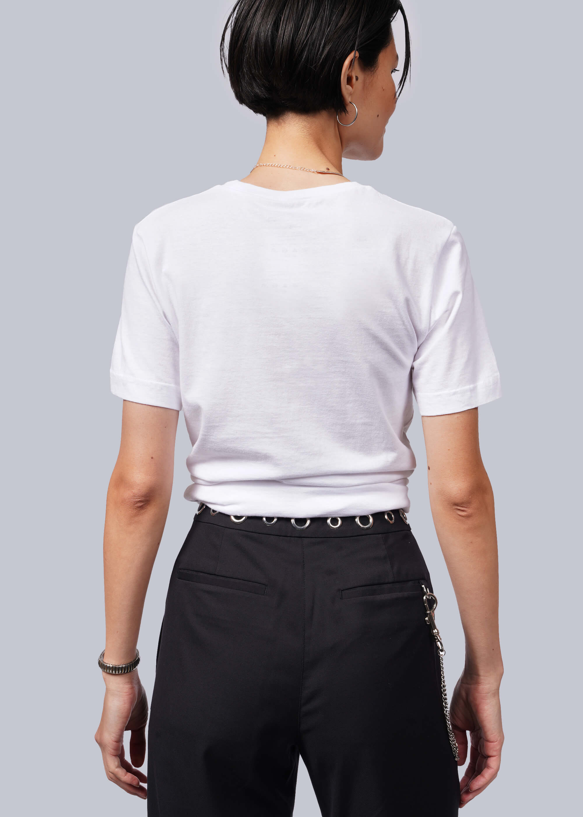 Rear view of a person with short dark hair in a relaxed-fit white The Wild Feminist® Tee and black pants featuring a silver chain on the side and waistband ring. The look is effortlessly stylish against a plain light gray background.
