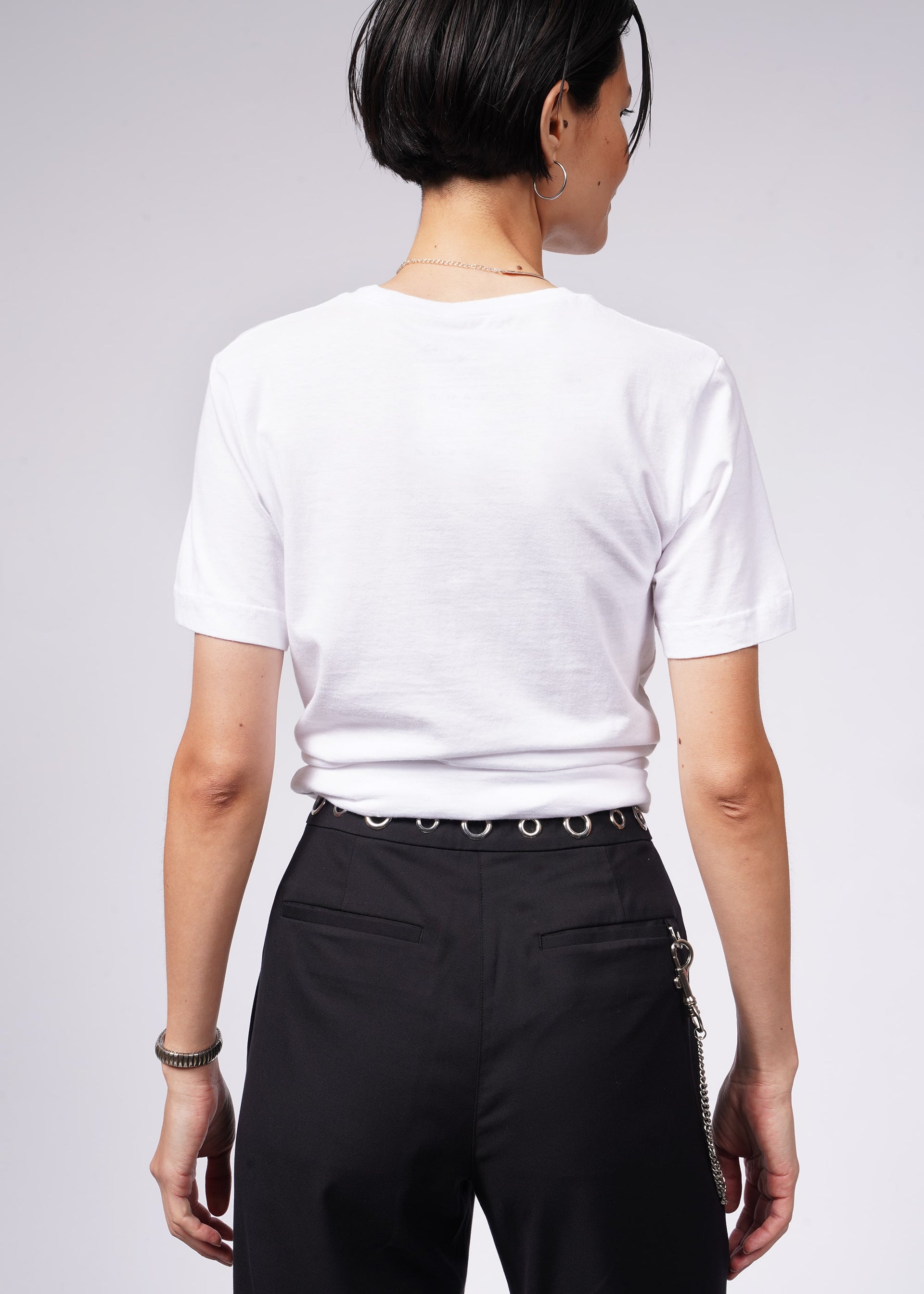 Rear view of a person with short dark hair in a relaxed-fit white The Wild Feminist® Tee and black pants featuring a silver chain on the side and waistband ring. The look is effortlessly stylish against a plain light gray background.