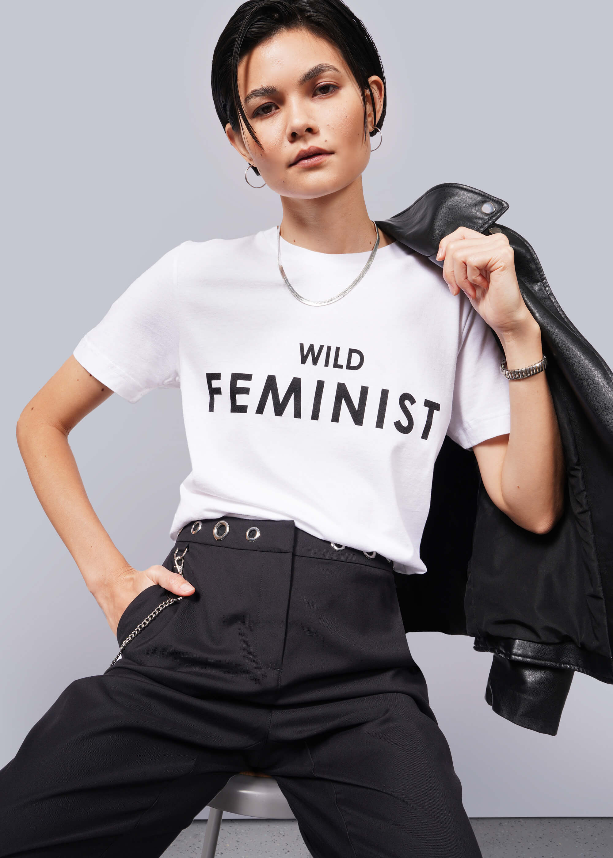 A person with short dark hair confidently wears The Wild Feminist® Tee with a relaxed, unisex fit. They hold a black leather jacket over their shoulder and pair it with high-waisted black pants, exuding modern empowerment against a plain backdrop.