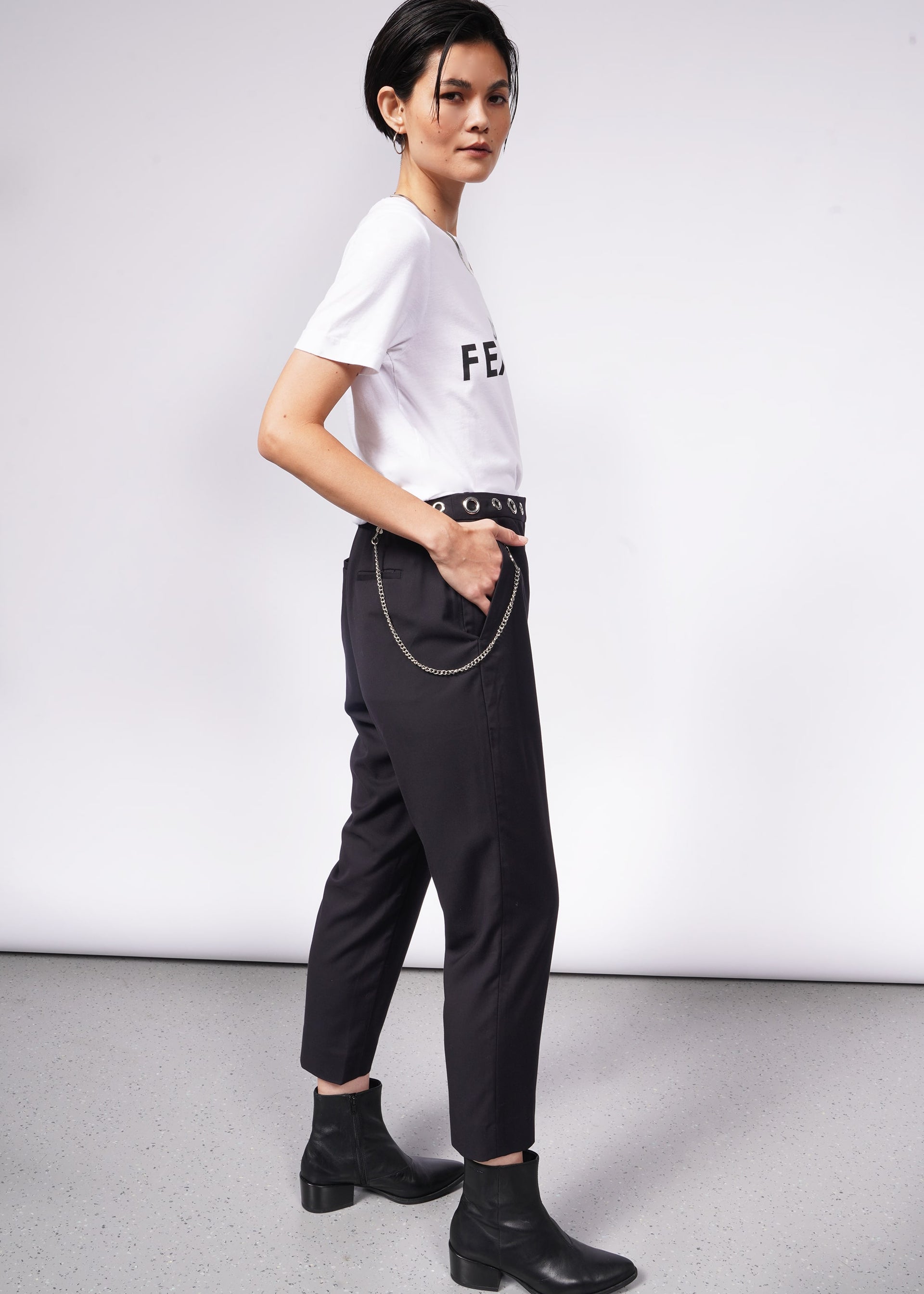 A person with short dark hair stands in profile against a white background, wearing The Wild Feminist® Tee—a relaxed unisex fit—paired with dark pants featuring a chain accessory and black boots. One hand rests casually in their pocket, exuding effortless style.
