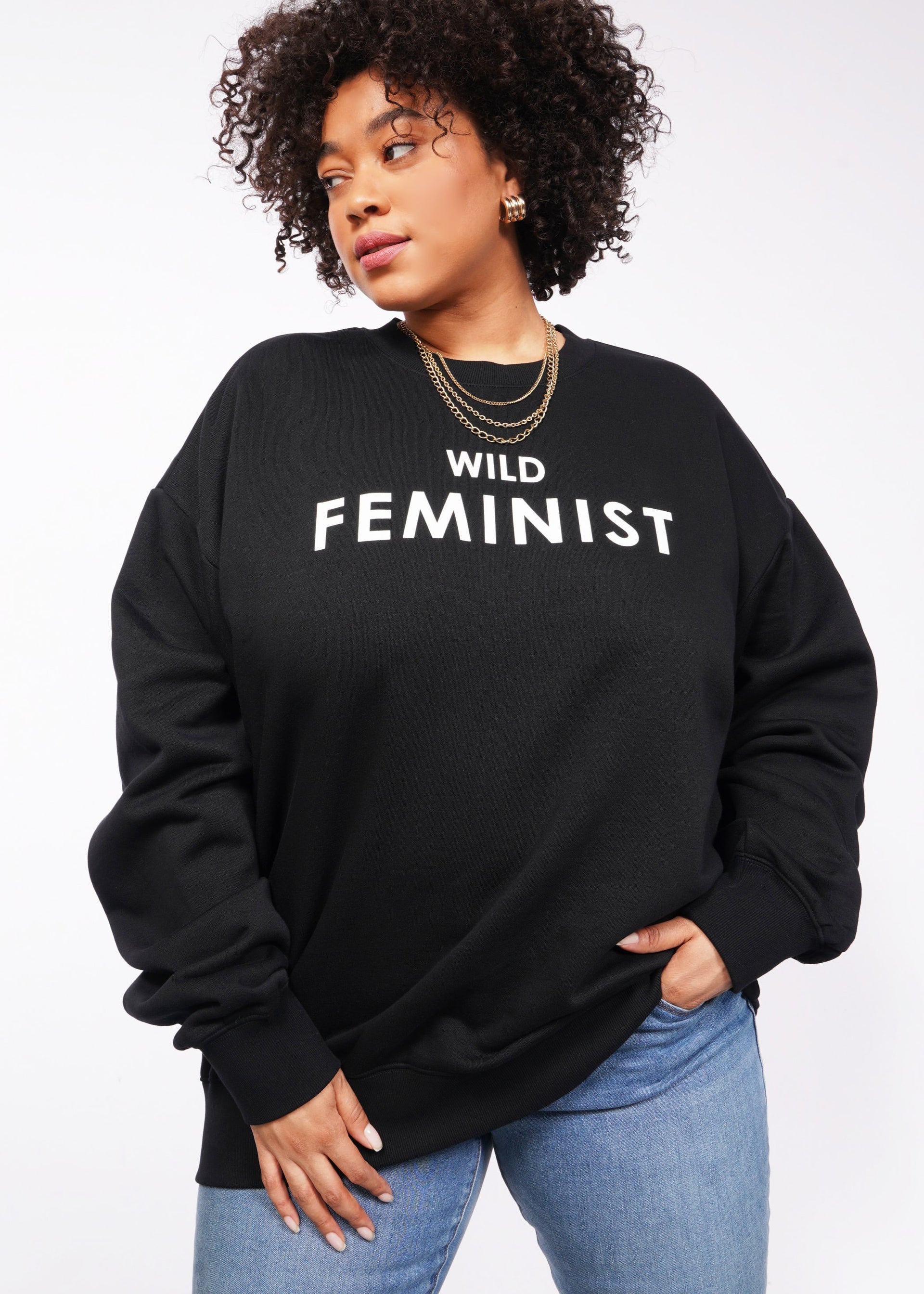 Against a plain white backdrop, someone sports the Wild Feminist Fleece Crew in black. The chic look is accentuated by bold WILD FEMINIST text, their curly hair, gold hoop earrings, and layered necklaces. The outfit is elevated by the crews soft brushed fleece feel.