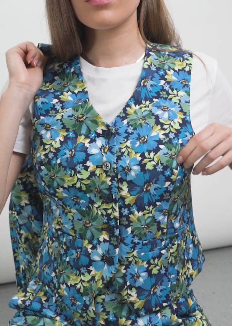 Video shows model with long blonde hair standing in front of white background wearing a 3-piece suit which includes The Empower Vest, The Empower Tux Blazer, and The Empower Wide Leg Pant in Carnation Navy, which is a blue toned floral print. The camera pans to a close up of the vest, its buttons, shows the model putting on the blazer, and then panning head to toe.