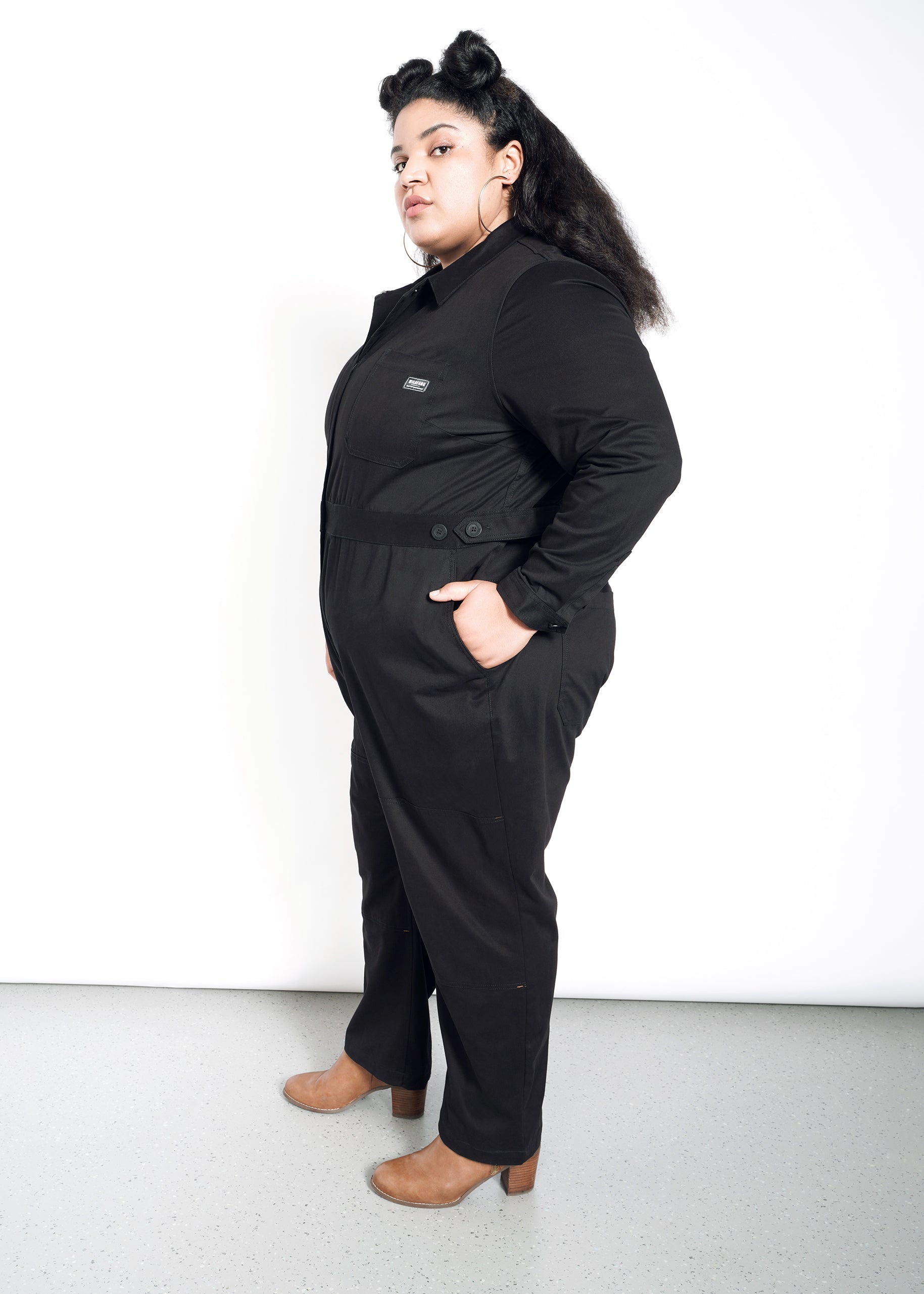 A person with long dark hair models The Essential Long Sleeve High Waisted Coverall in black, featuring an adjustable waist and deep pockets, paired with tan shoes, standing sideways against a plain white background.