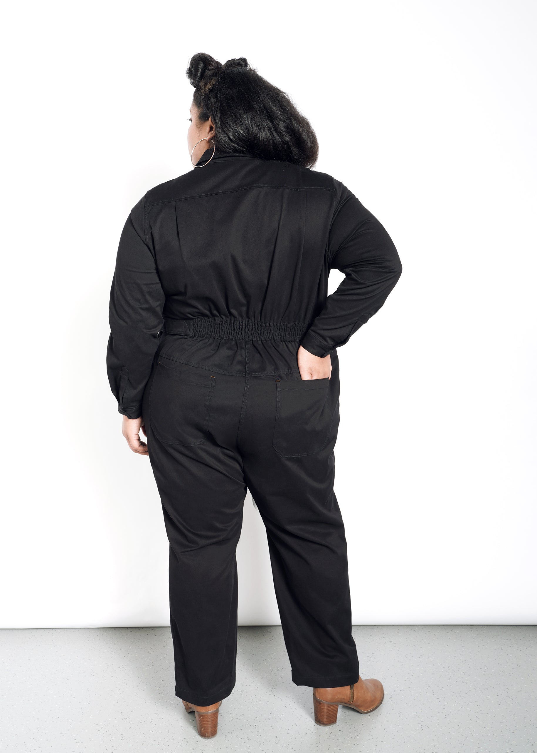 A person with long dark hair stands facing away from the camera against a white background, wearing The Essential Long Sleeve High Waisted Coverall in black with deep pockets and brown heeled shoes. One hand rests on their hip.