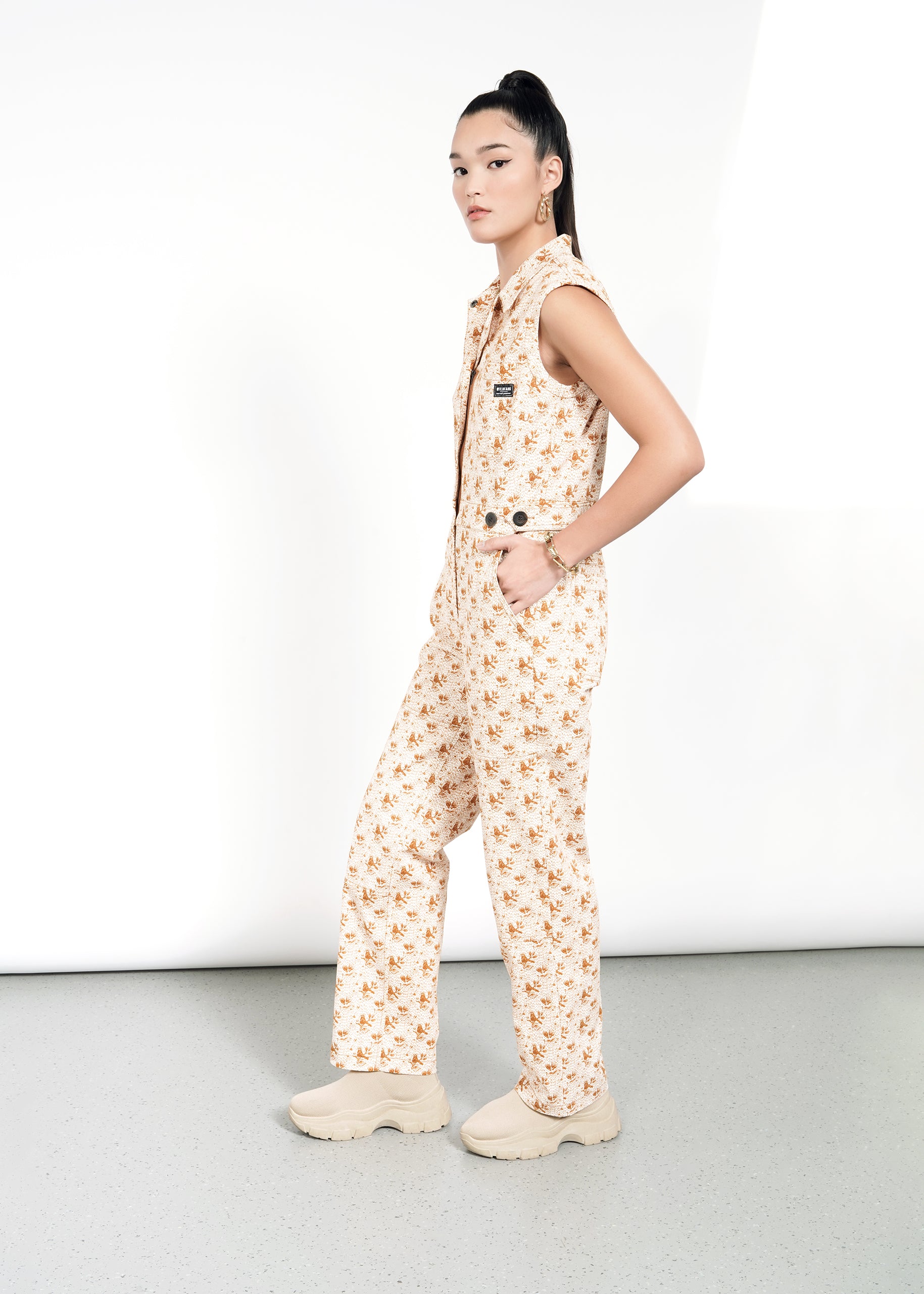 A woman with her hair in a high ponytail stands sideways against a light background, wearing The Essential Sleeveless High Waisted Coverall— a light-colored, floral-patterned jumpsuit featuring deep pockets.