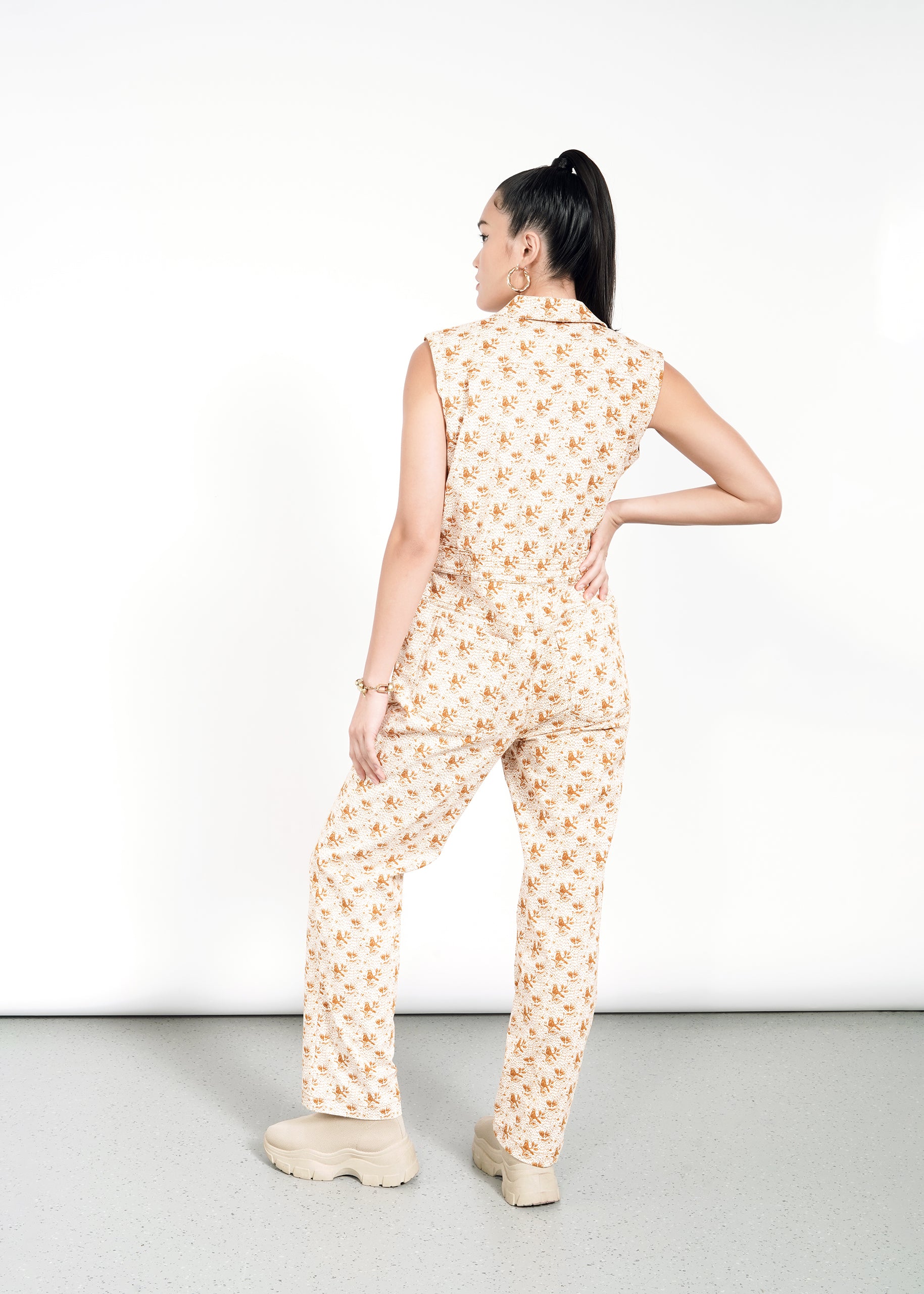 A woman stands with her back to the camera, one hand on her hip, wearing The Essential Sleeveless High Waisted Coverall with a floral print and chunky beige shoes. Her hair is in a high ponytail against a plain white background.