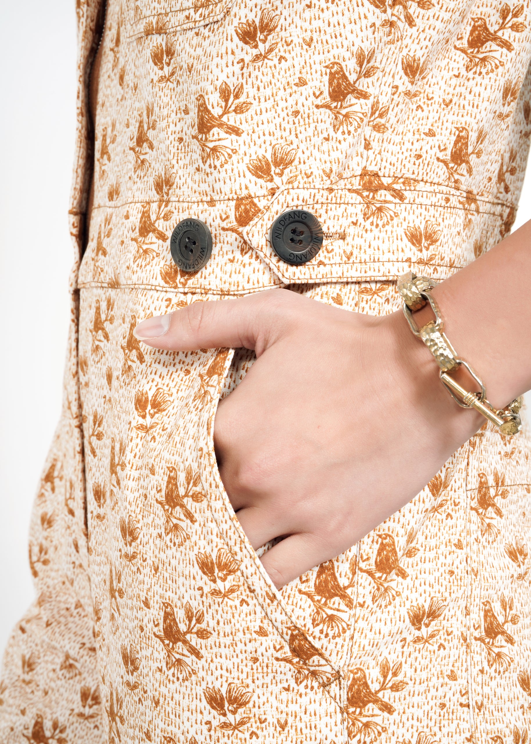 A close-up of a hand in the deep pocket of The Essential Sleeveless High Waisted Coverall, a beige outfit with brown bird and leaf patterns, adjustable waist, two black buttons, and a gold chain bracelet on the wrist.