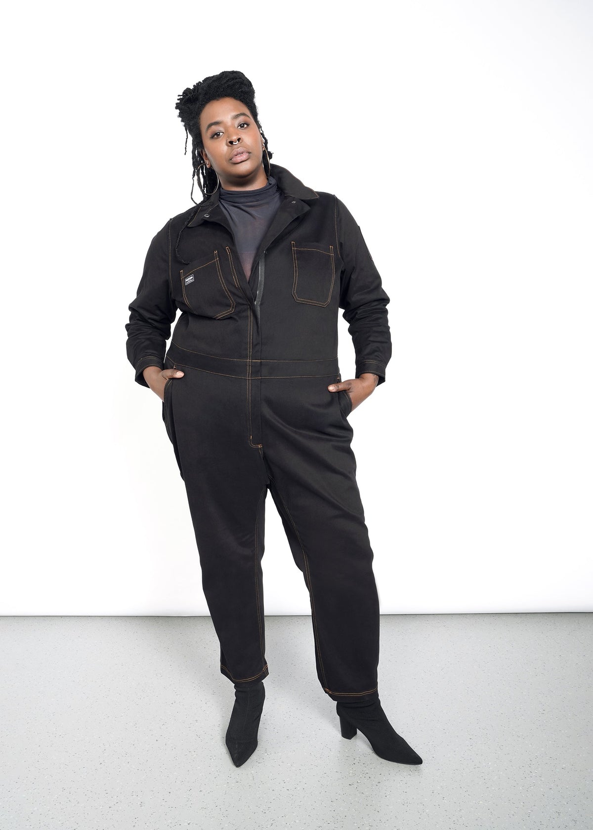 model wearing black cotton stretch coverall jumpsuit boiler suit long sleeve with brown spice colored pop stitching. pants are full length