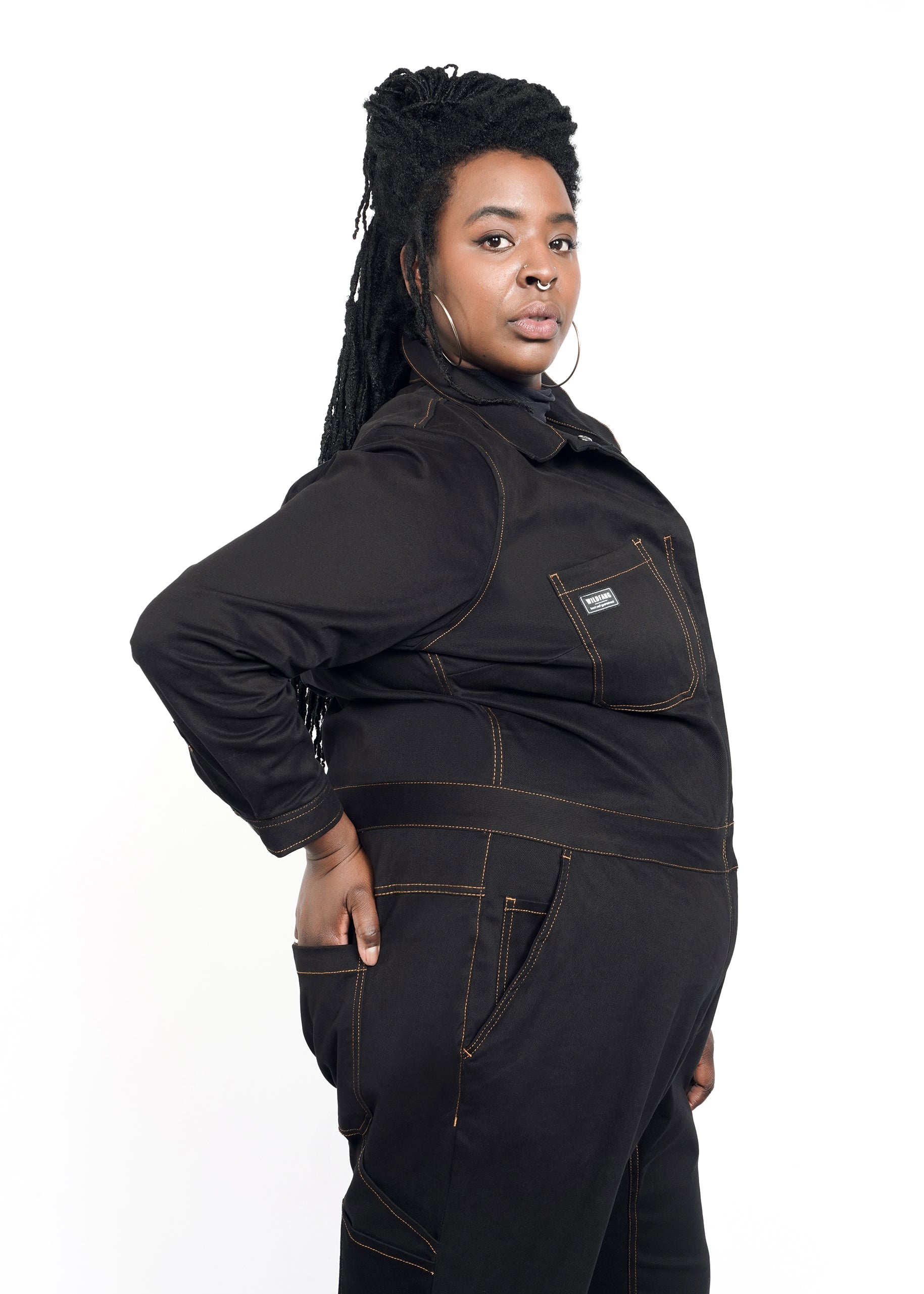 Model wearing black size small long sleeve coverall jumpsuit with orange pop-stitch detailing, sitting on stool. Sleeves and pant legs are cuffed