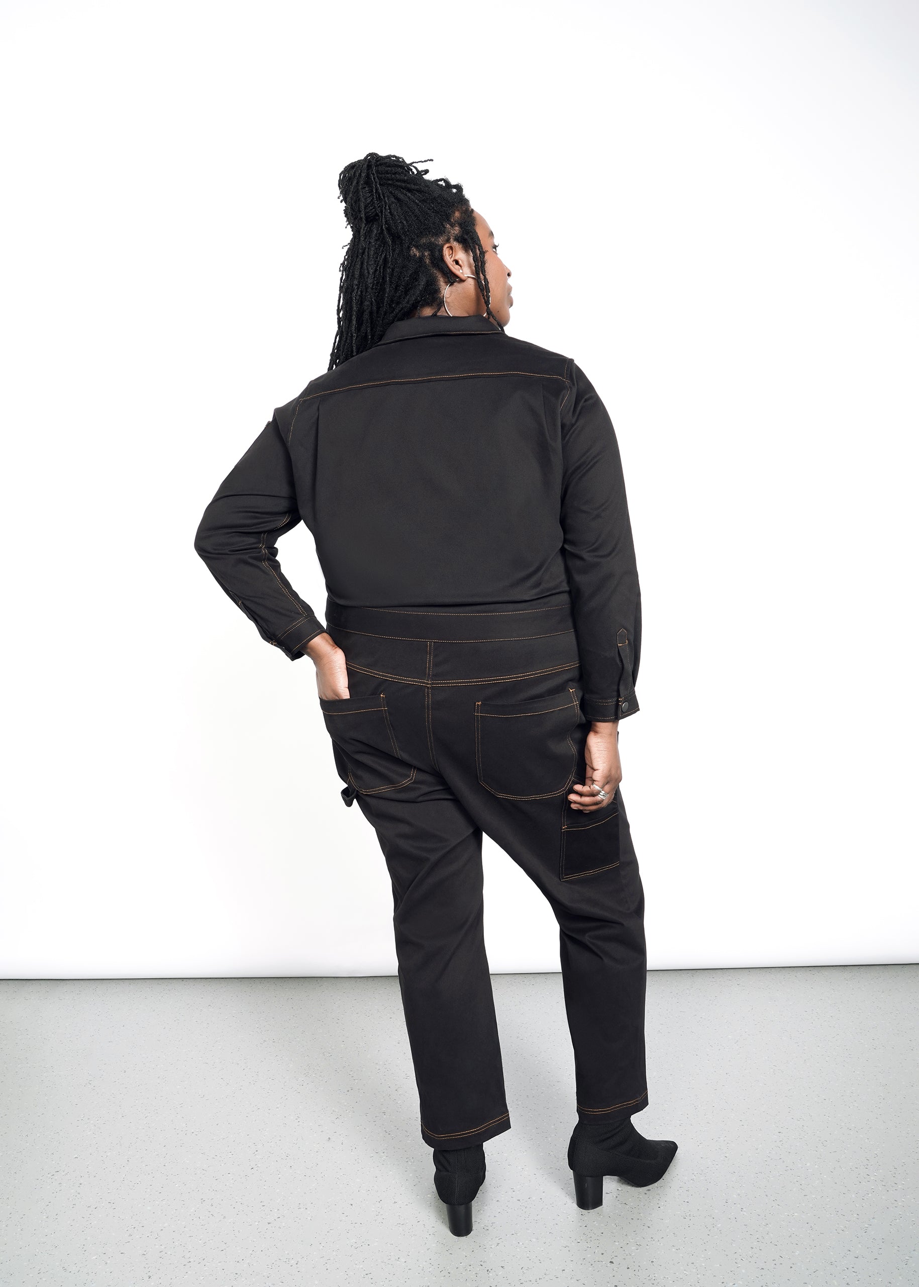 model wearing black cotton stretch coverall jumpsuit boiler suit long sleeve with brown spice colored pop stitching. pants are full length