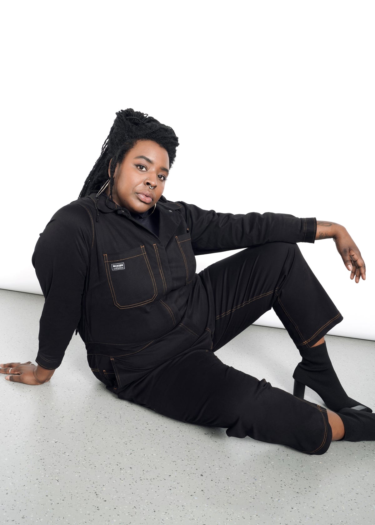 model wearing black cotton stretch coverall jumpsuit boiler suit long sleeve with brown spice colored pop stitching. pants are full length