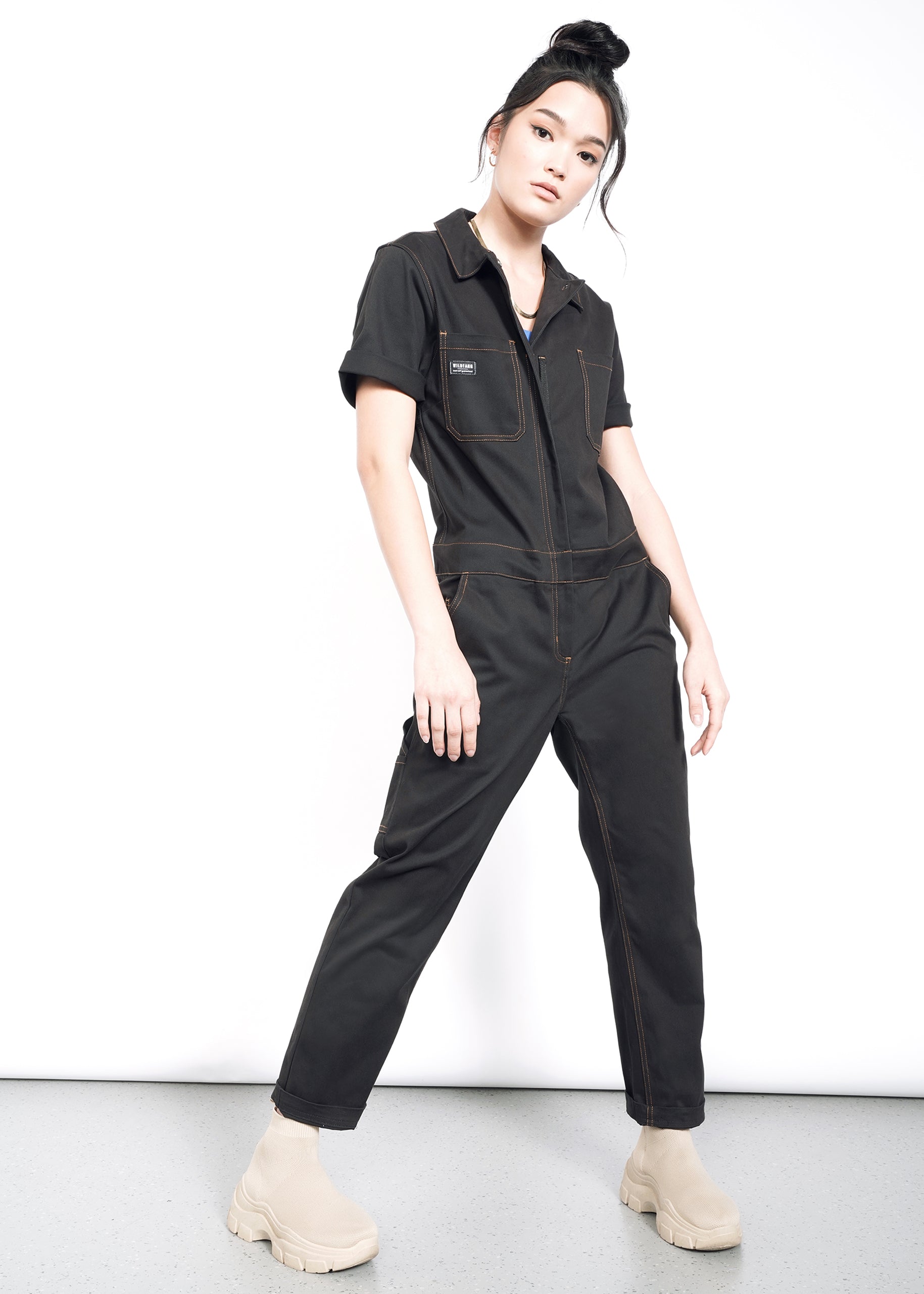 A woman poses confidently against a white background, wearing The Essential Coverall—a black, short-sleeve jumpsuit with multiple pockets—paired with beige boots. Her dark hair is in a top bun as she looks at the camera, hand on her thigh.