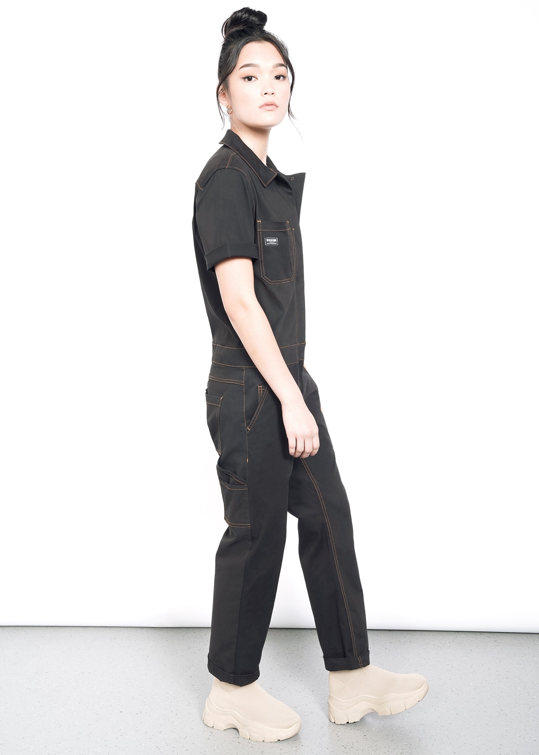 A person with long black hair in a topknot wears The Essential Coverall—a black jumpsuit with visible stitching and multiple pockets—paired with beige chunky boots, standing sideways against a plain white background.