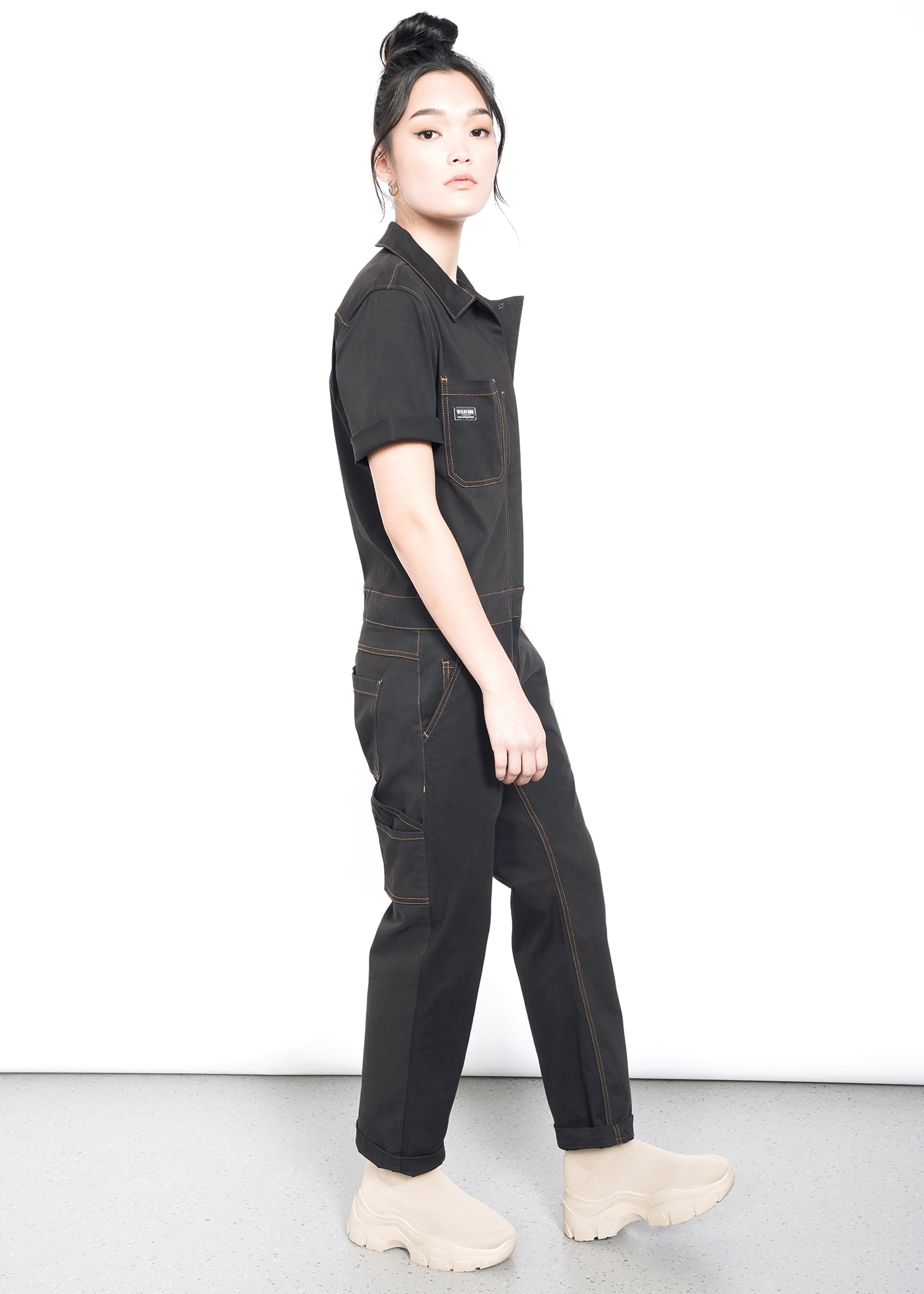 model wearing black cotton stretch coverall jumpsuit boiler suit short sleeve with brown spice colored pop stitching