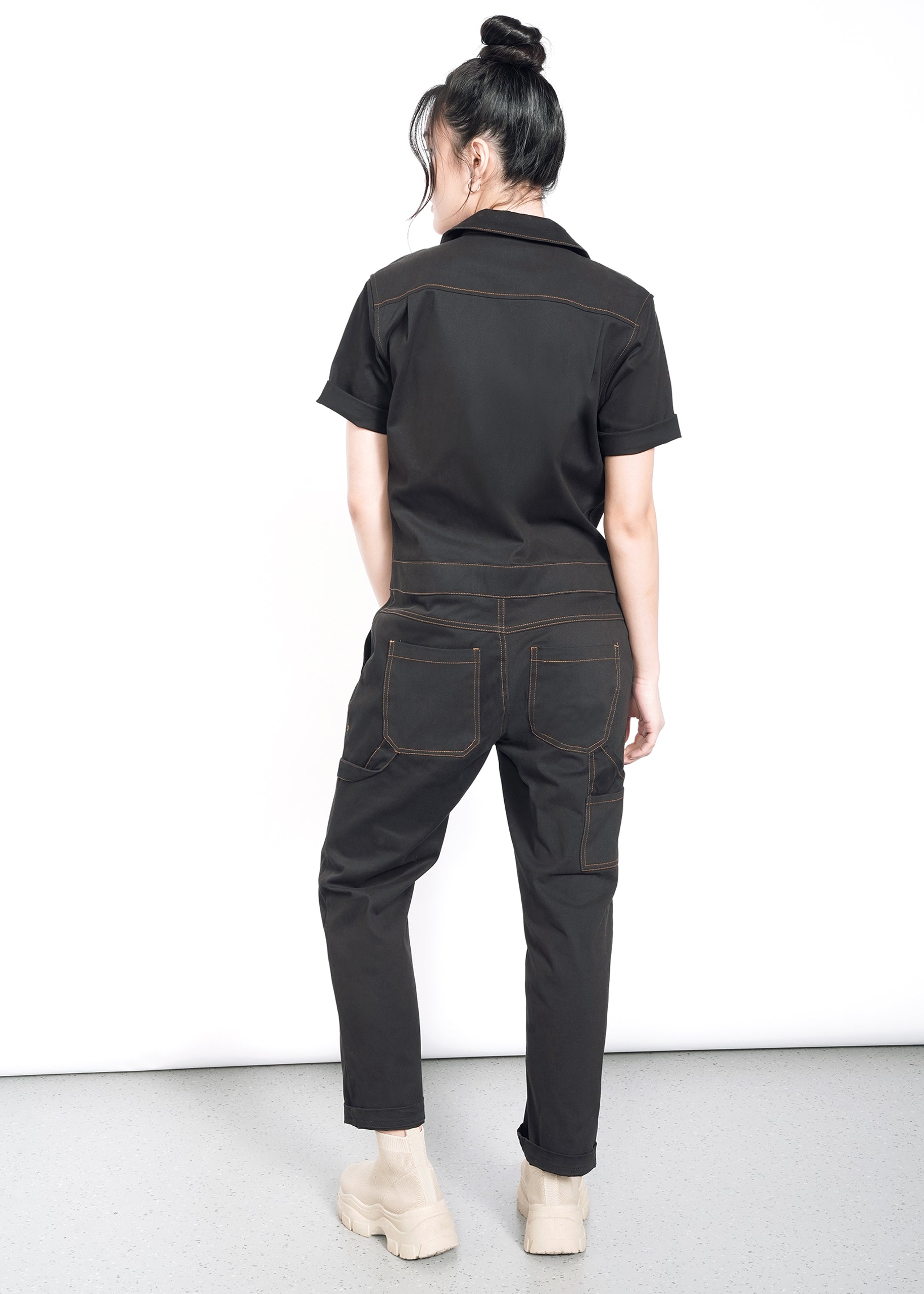 A person with long dark hair in a bun wears The Essential Coverall, a black stretch cotton short-sleeve jumpsuit with contrast stitching, and beige boots, standing with their back to the camera against a plain white background.