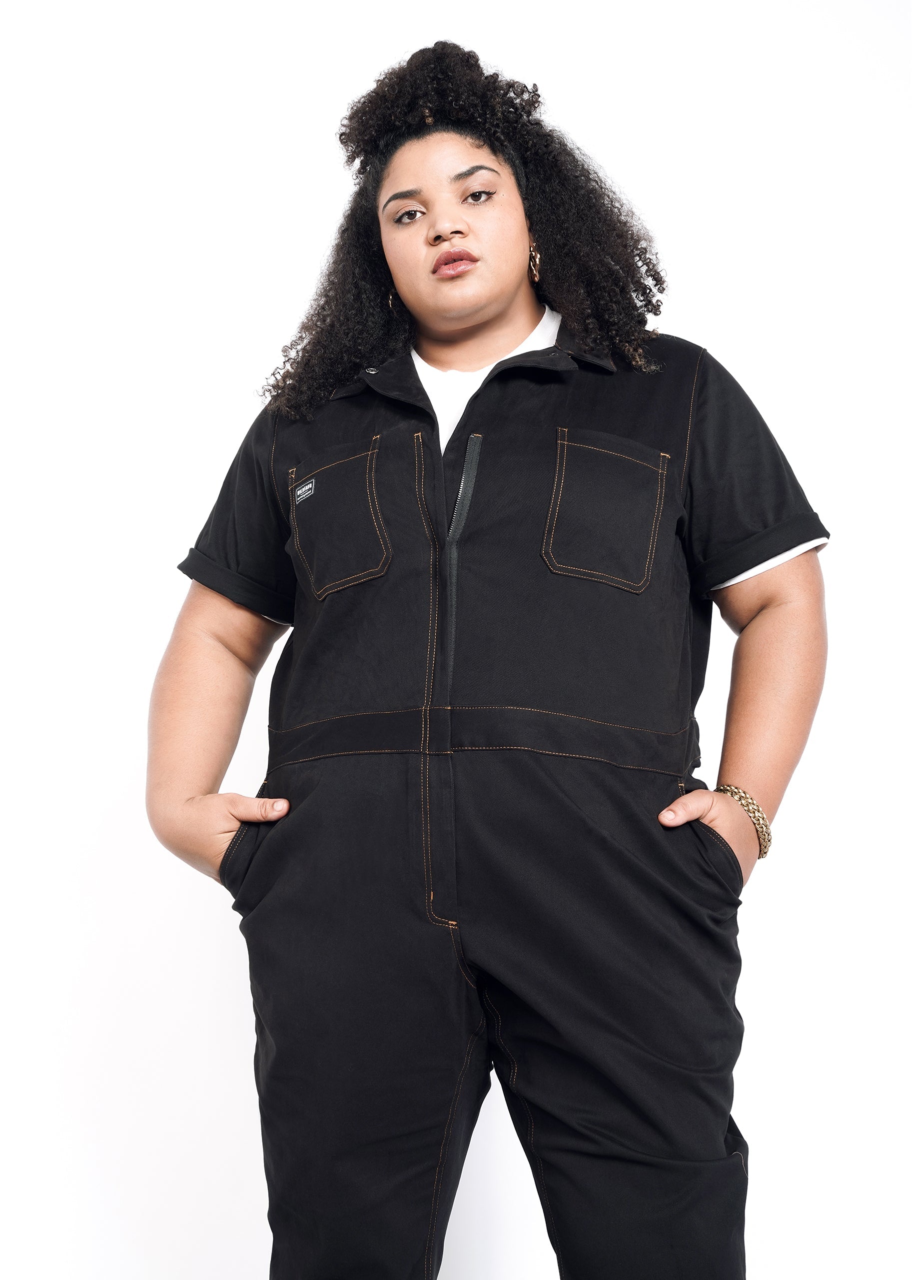 A person with curly hair stands confidently against a white background, wearing The Essential Coverall—a black jumpsuit crafted from soft brushed stretch cotton with visible stitching and hands in pockets.