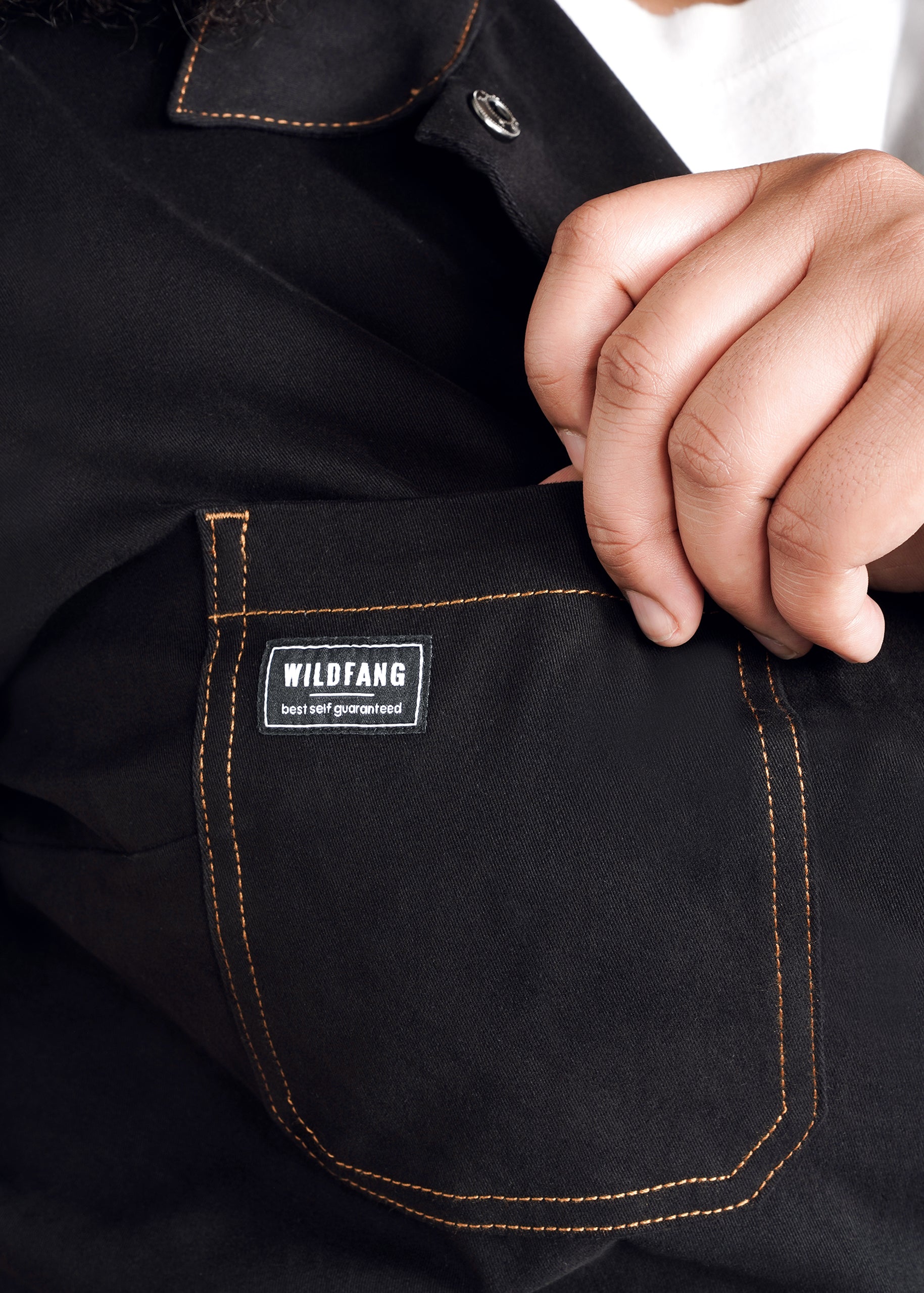 A close-up of a hand holding the edge of The Essential Coverall’s black pocket, crafted from soft brushed stretch cotton with orange stitching and a “WILDFANG best self guaranteed” tag—one of 9 pockets on the garment.
