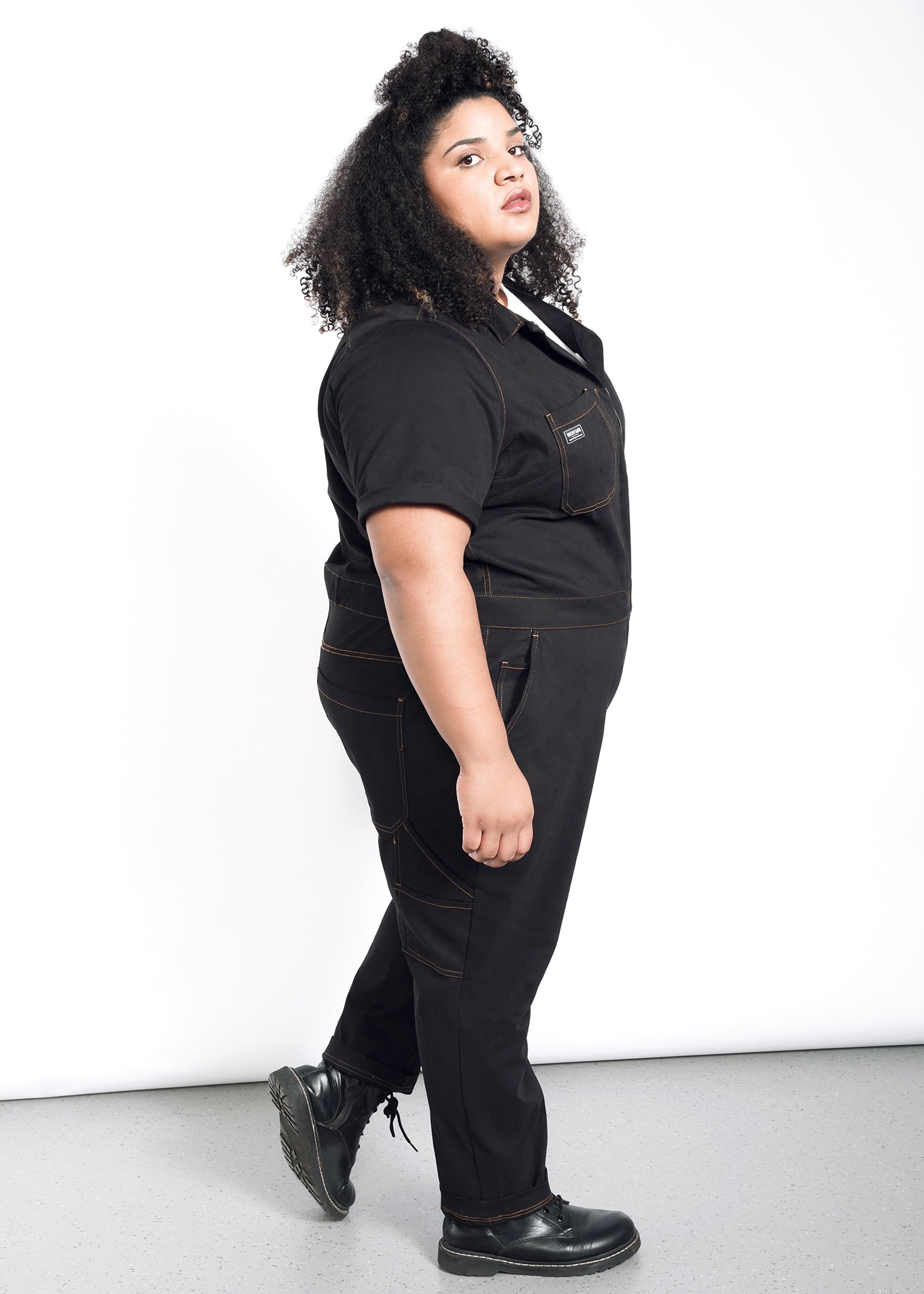 A person with curly hair stands sideways, looking at the camera. They wear The Essential Coverall in black—crafted from soft brushed stretch cotton with 9 pockets and brown stitching—paired with a black t-shirt and shoes against a white background.