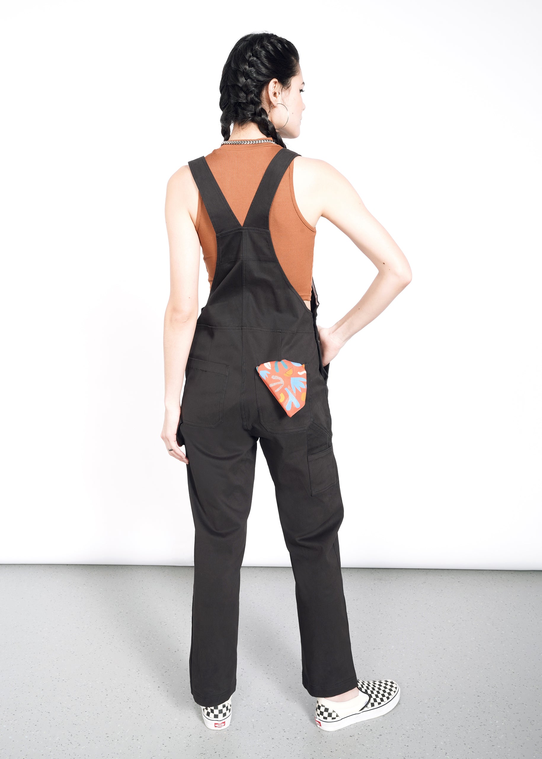 A person with braided hair faces away from the camera, wearing The Essential Overall in black with adjustable straps, deep pockets, and an orange patch with a blue pattern. Theyre paired with a rust sleeveless top and checkered slip-on shoes made of soft stretch cotton.