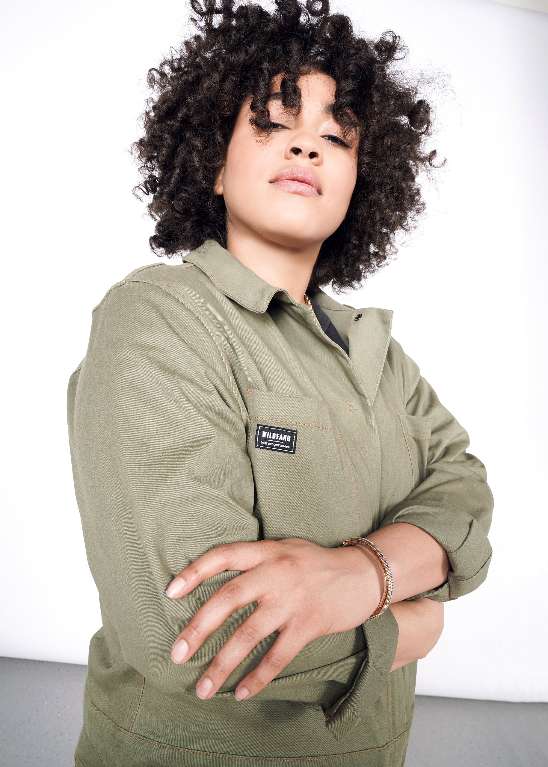 Curly haired model wearing olive long sleeve coveralls with arms folded at chest