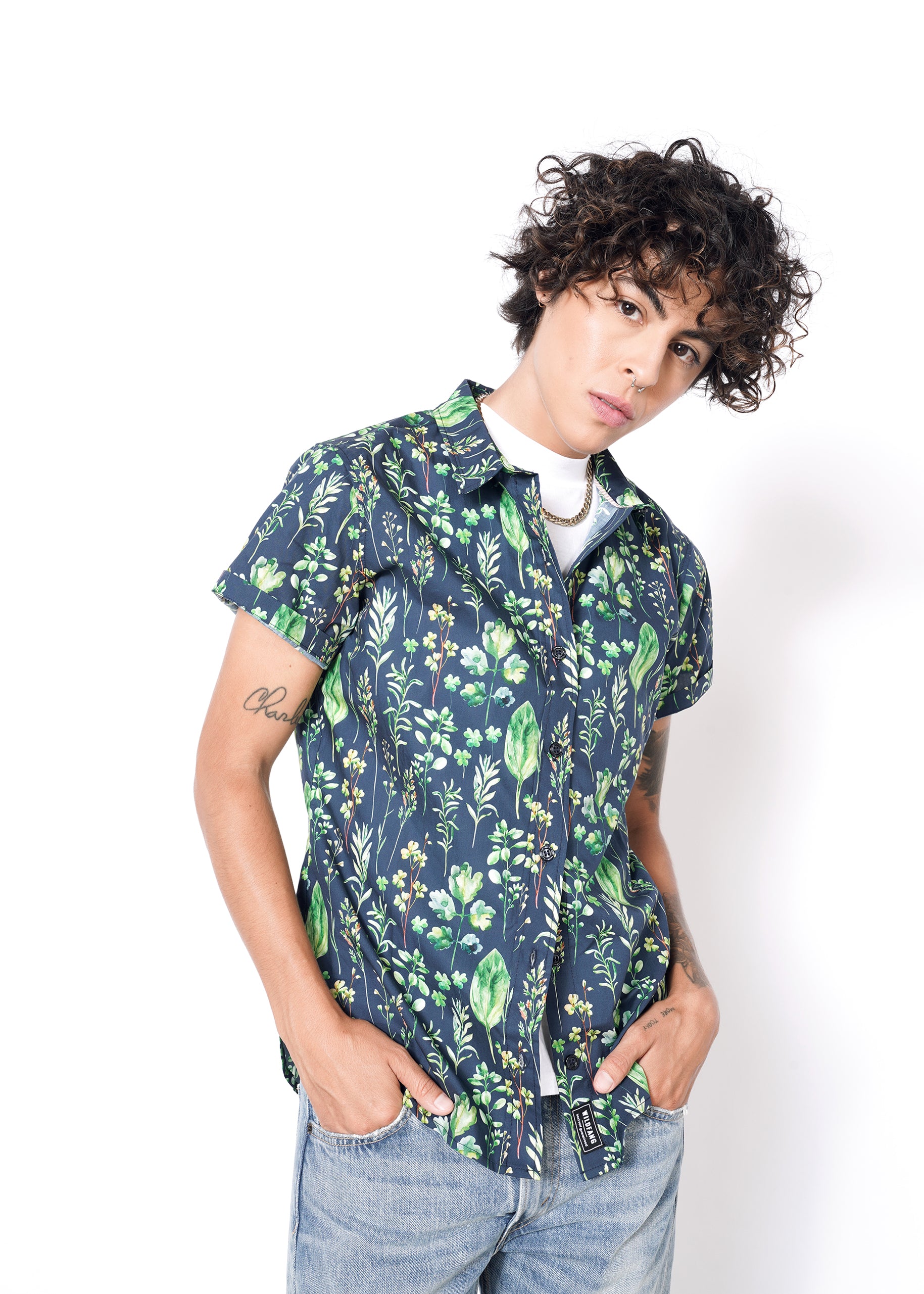 A person with curly hair confidently wears The Essential Button Up in navy with green floral patterns, tailored fit, and short sleeves. Underneath is a white shirt, paired with light blue jeans against a crisp plain white background. Hands are casually tucked in pockets.