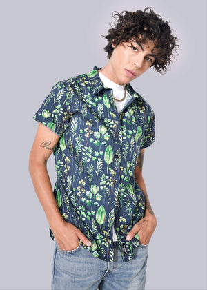 S= A person with curly hair confidently wears The Essential Button Up in navy with green floral patterns, tailored fit, and short sleeves. Underneath is a white shirt, paired with light blue jeans against a crisp plain white background. Hands are casually tucked in pockets.
