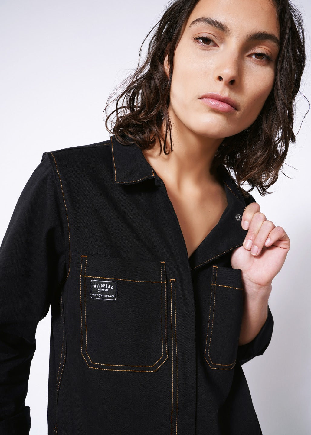 Model wearing black size small long sleeve coverall jumpsuit with orange pop-stitch detailing, pulling on collar and looking into camera