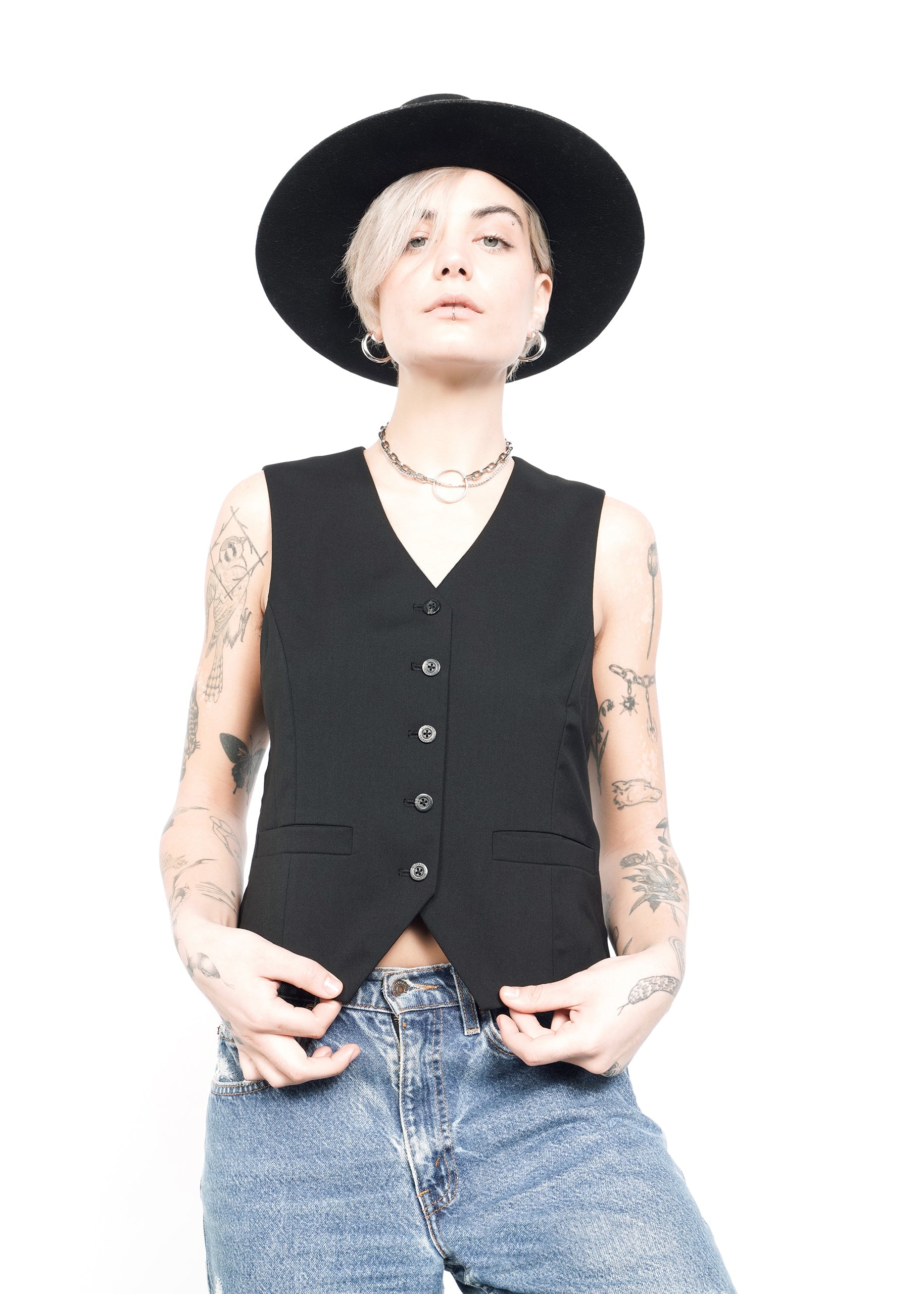 A person with a short haircut wears The Empower Vest, a tailored fit, over blue jeans and tops it off with a black wide-brimmed hat. Tattooed arms hold the sides of the vest against a plain white background.