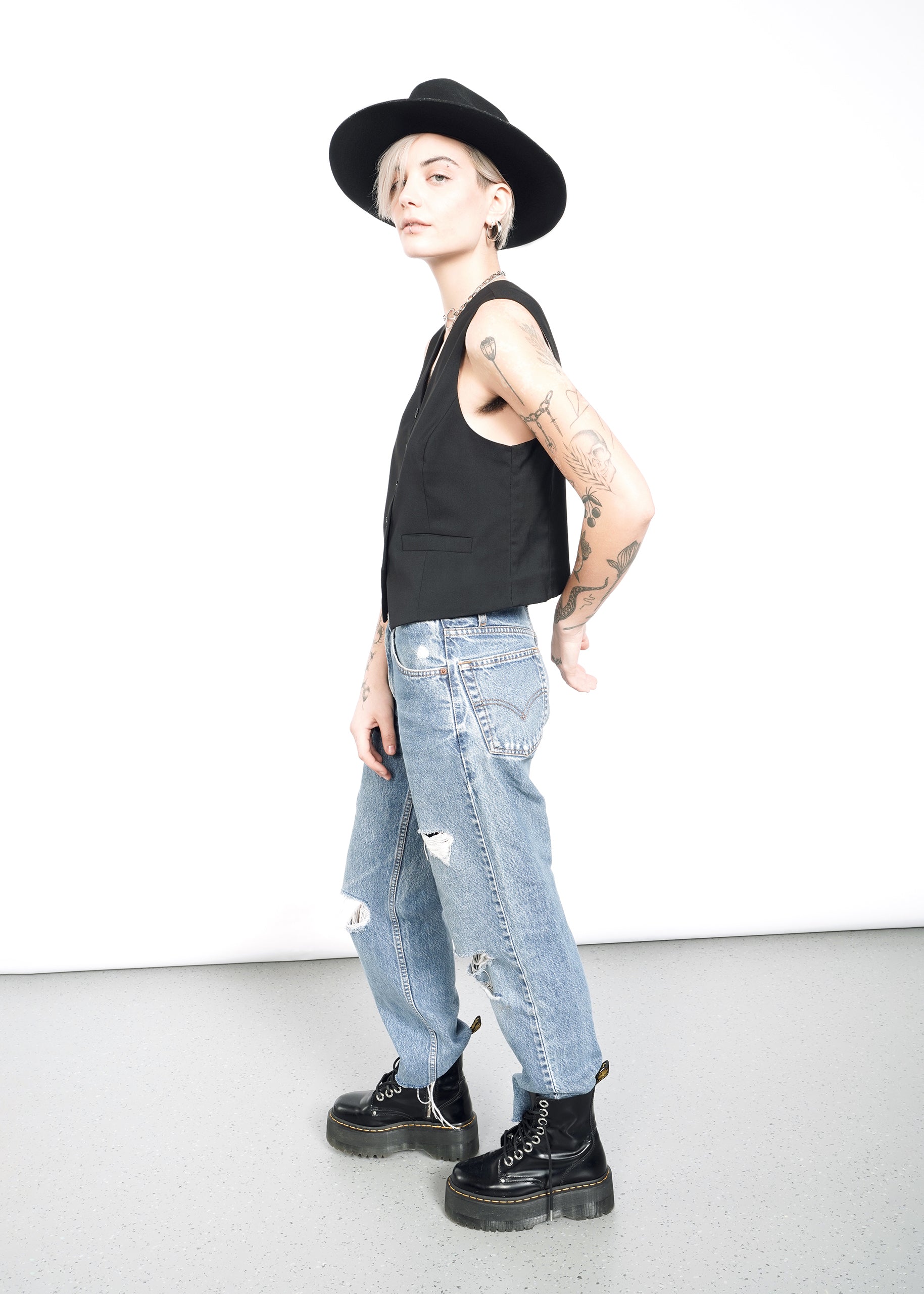 A person stands sideways in a tailored fit black hat, ripped jeans, and The Empower Vest, showcasing sleeveless layers. Tattoos adorn their arms, paired with black boots. The plain light-colored wall and floor backdrop highlight the ensembles edgy style.