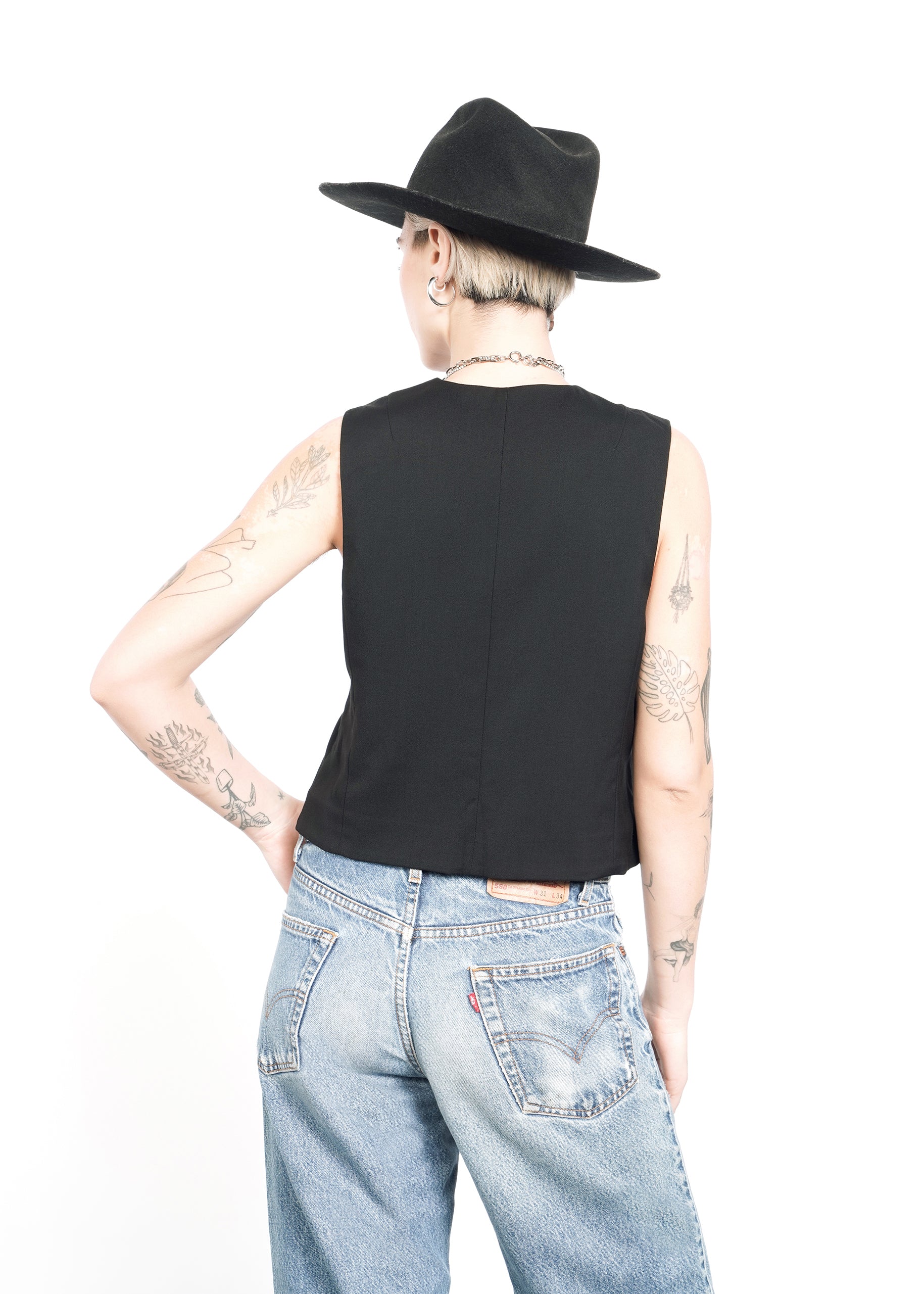 A person in a black sleeveless top expertly layered over The Empower Vest pairs it with light blue jeans and a black hat. Facing away, they showcase short hair and arm tattoos against a plain white backdrop.