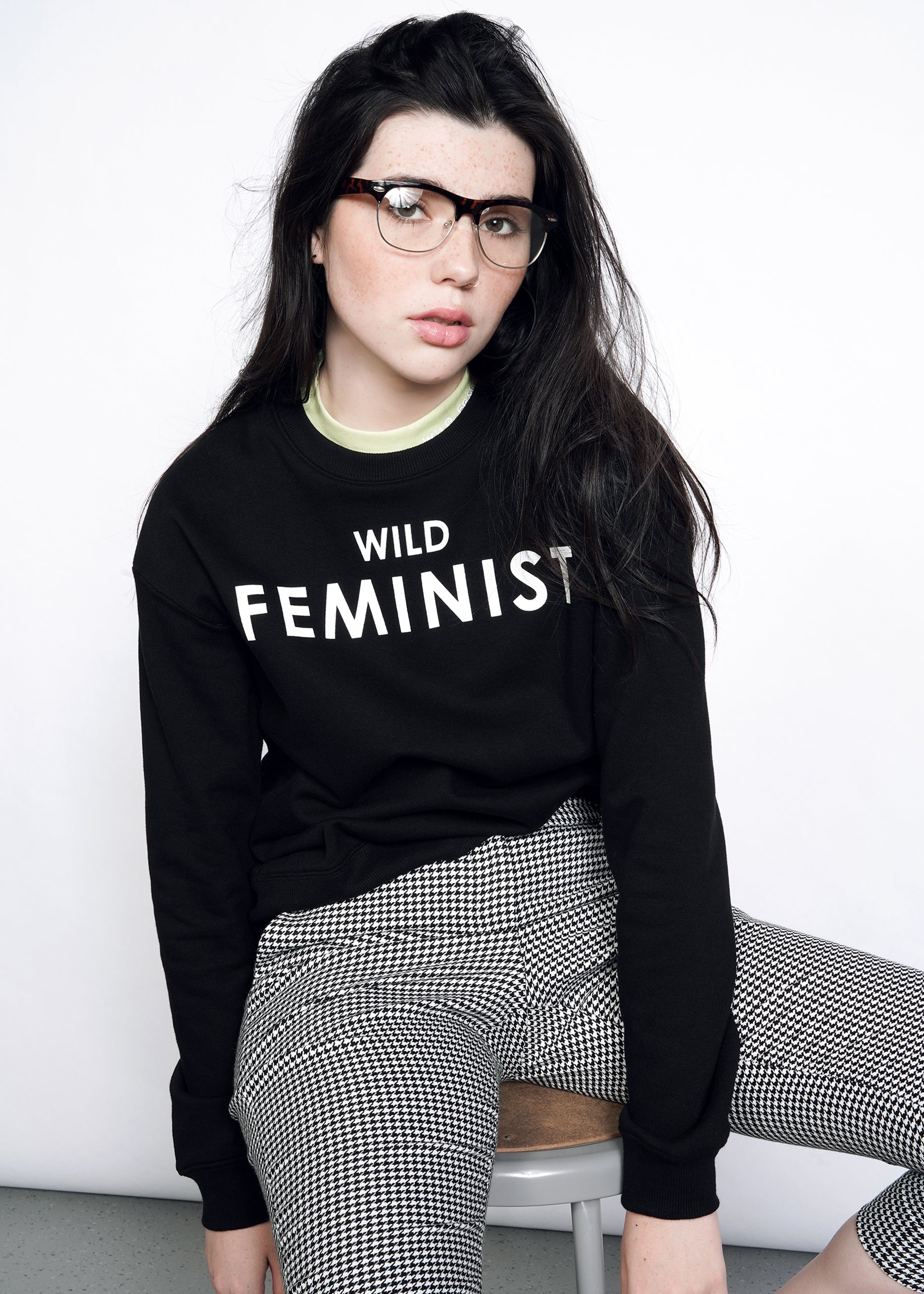 Model with dark hair wearing black sweatshirt that reads Wild Feminist in white, styled with glasses, green undershirt, and houndstooth pants.