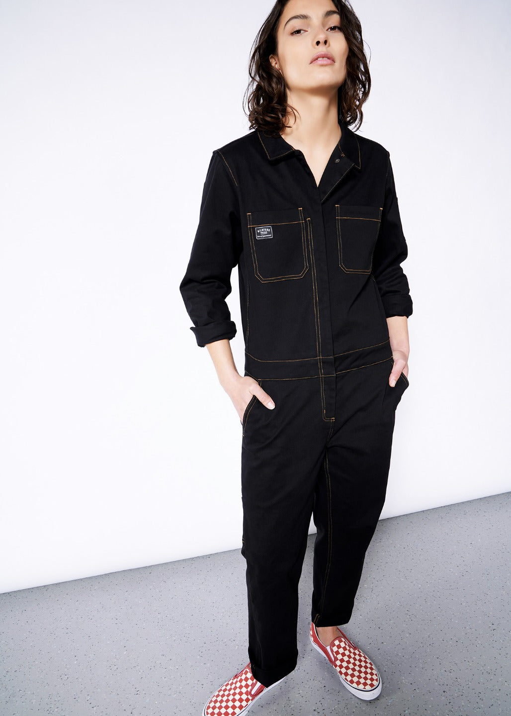 Model standing with hands in pockets, wearing black size small long sleeve coverall jumpsuit with orange pop-stitch detailing. Pant legs are rolled down and top button is un buttoned.