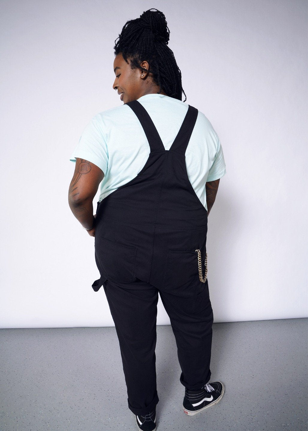 Backside of model wearing black overalls with metal hardware in size XXL, with an blue tee underneath and chain in back pocket