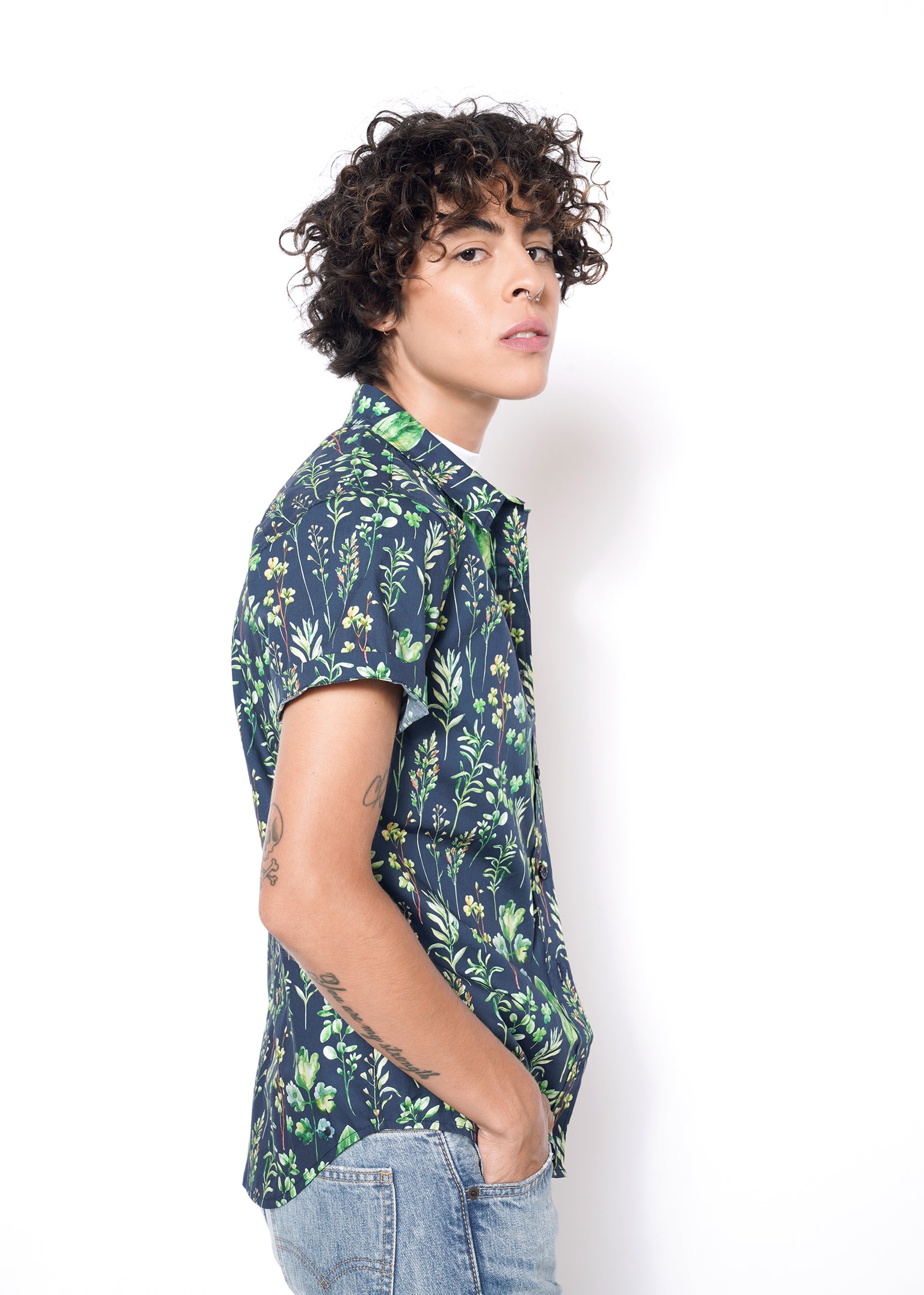With curly hair, theyre wearing The Essential Button Up with short sleeves in blue featuring a green floral pattern in a tailored fit that enhances their style. Paired with light blue jeans, they pose slightly turned toward the camera against a plain white background.