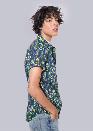 S= With curly hair, theyre wearing The Essential Button Up with short sleeves in blue featuring a green floral pattern in a tailored fit that enhances their style. Paired with light blue jeans, they pose slightly turned toward the camera against a plain white background.