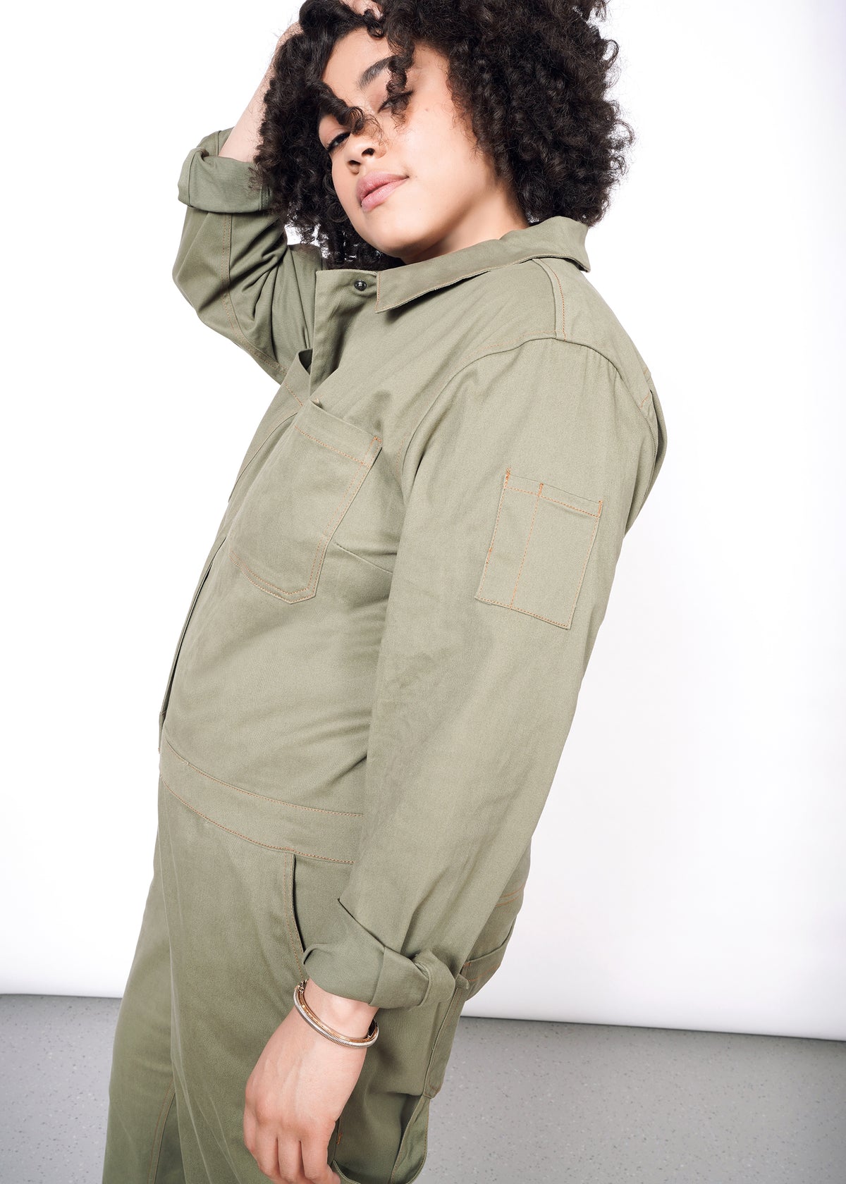 Profile view of curly haired model wearing olive long sleeve coveralls, showing the pocked and orange stitching on sleeve
