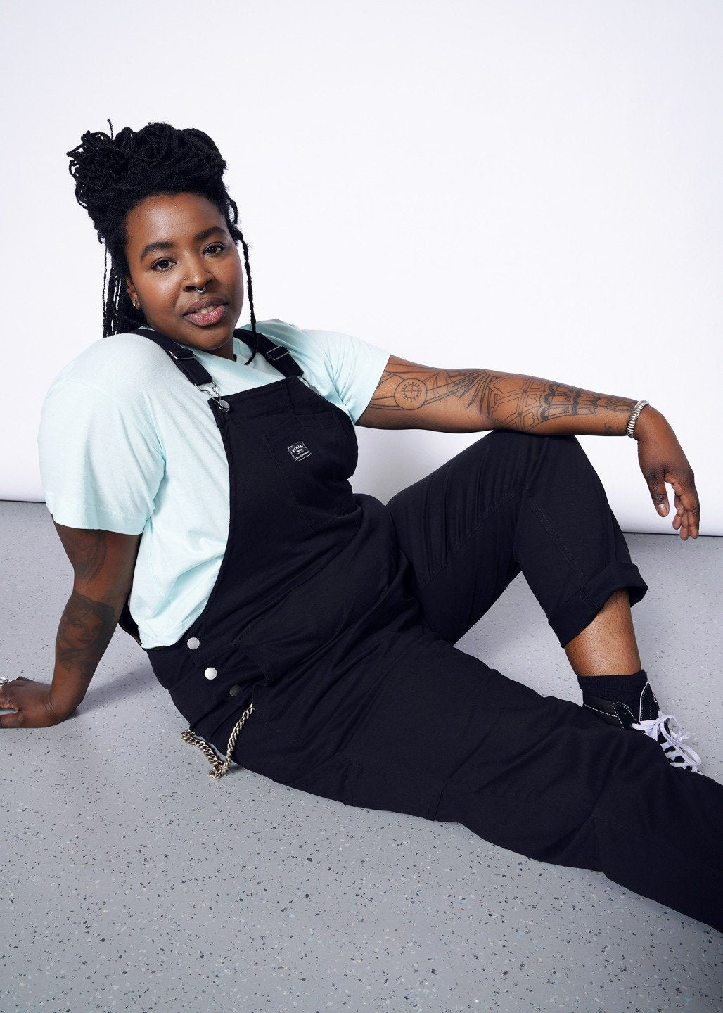 Model wearing black overalls with metal hardware in size XXL, with an blue tee underneath, sitting on the floor with one leg bent
