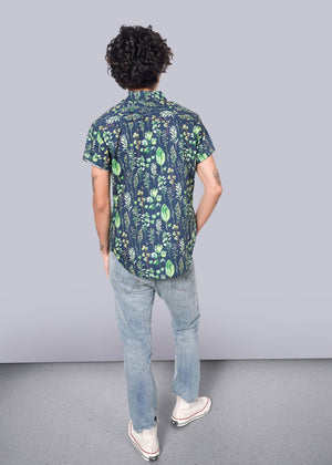 S=A person with curly hair, seen from the back, wears The Essential Button Up, a short-sleeved blue shirt with a green leaf pattern made of 100% cotton. They pair it with light blue jeans and white sneakers while standing in front of a plain white wall on a gray floor.