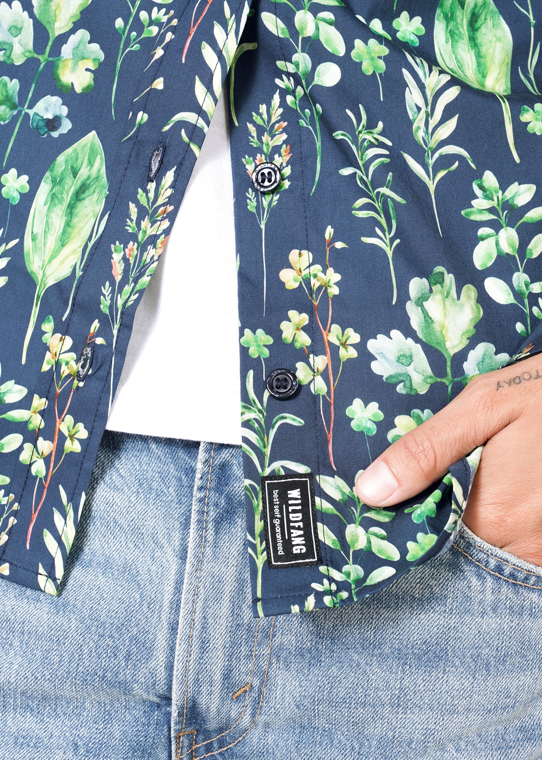 A person is wearing The Essential Button Up—a dark blue, short-sleeved 100% cotton shirt with a green leaf and botanical print. The shirt is partly buttoned over a white top, paired with light blue jeans. Their tattooed hands are visible. A clothing label reads WILDLIING.